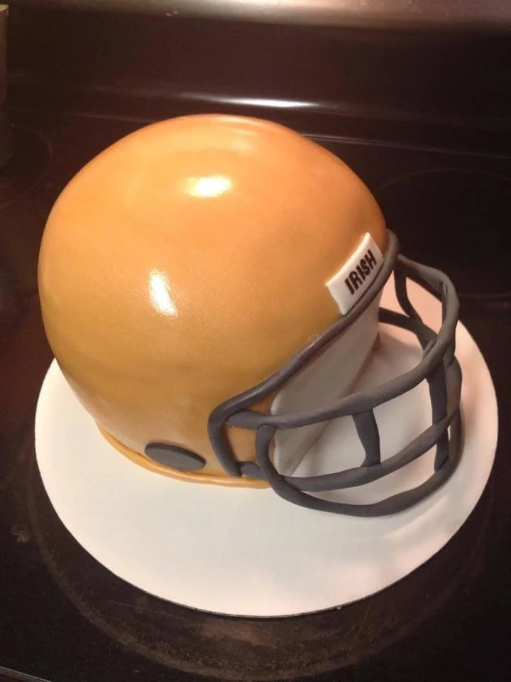 A cake designed to look like a football helmet with a gold top, black face mask, and a fortune sticker labeled 'IRISH.'