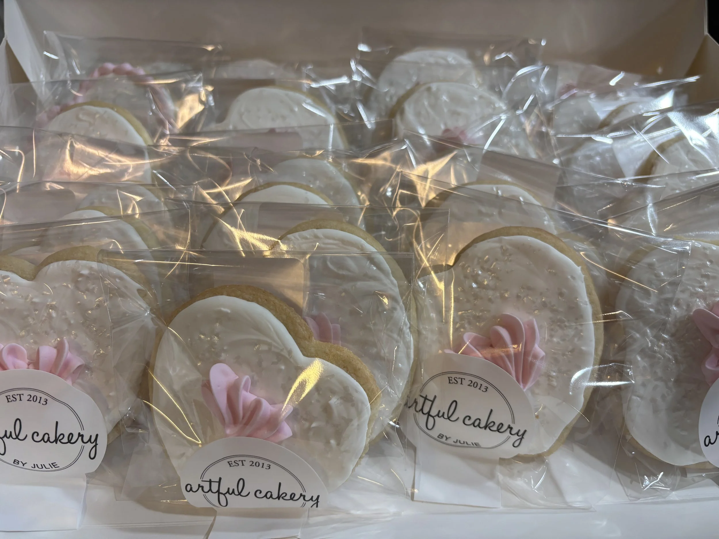 Set of heart-shaped cookies decorated with white icing and pink icing flower accents, individually wrapped in clear plastic bags with labels that read "artful cakery BY JULIE," inside a white box.