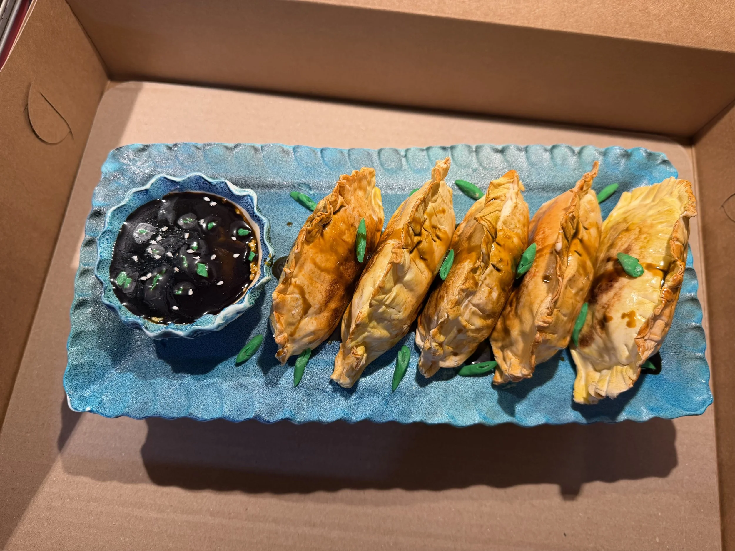 Overhead view of a realistic fried dumpling cake with five gyoza, edible dipping sauce bowl, and handmade blue edible platter.