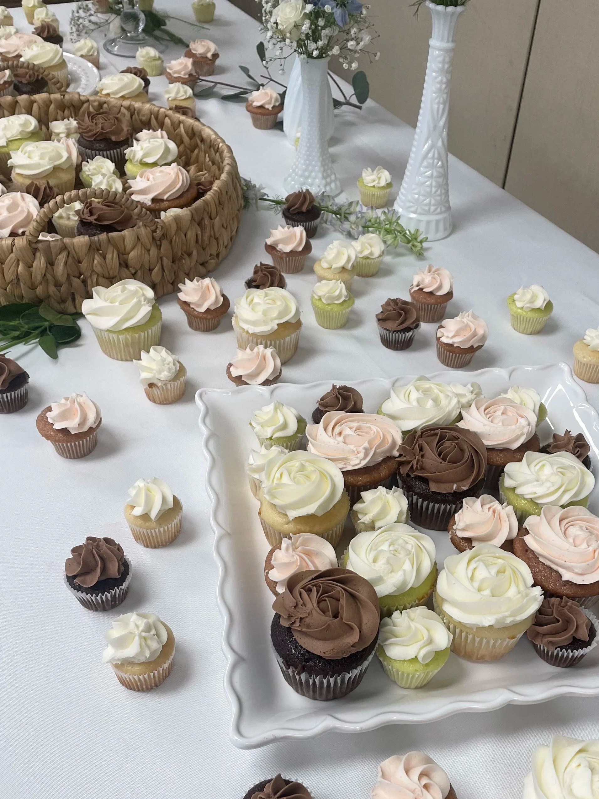 Assorted cupcakes with swirled frosting in white, pink, and brown, displayed on a white table with decorative vases and a woven basket.