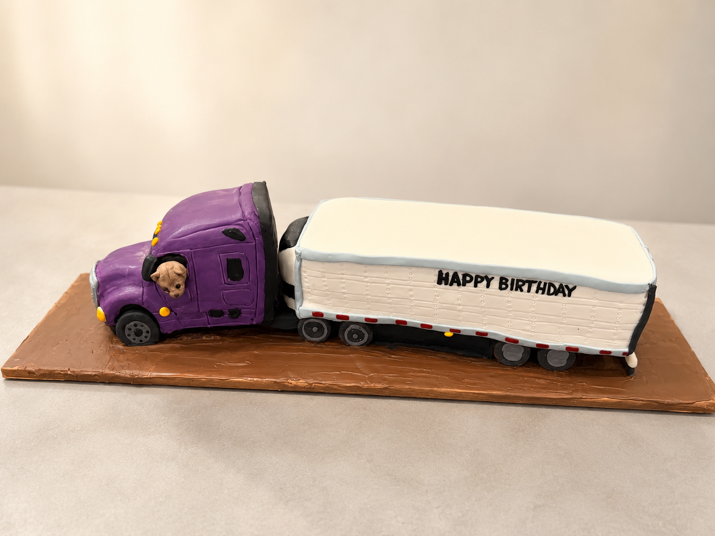 A custom semi truck and trailer birthday cake, handcrafted from cake and decorated with hand-painted details. Complete with a sweet dog peeking out the driver's window, this realistic truck cake was designed for a trucking enthusiast's special day.