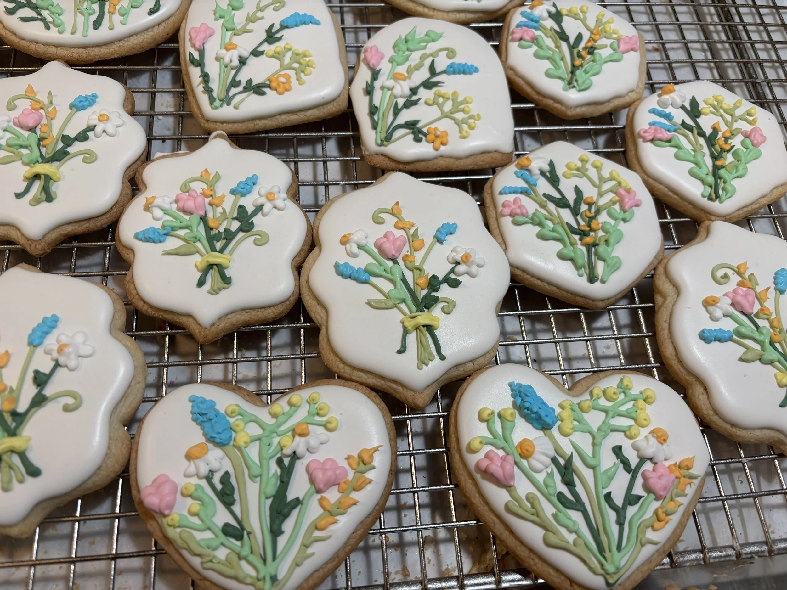 Heart-shaped cookies decorated with white icing and colorful floral designs, placed on a wire cooling rack.