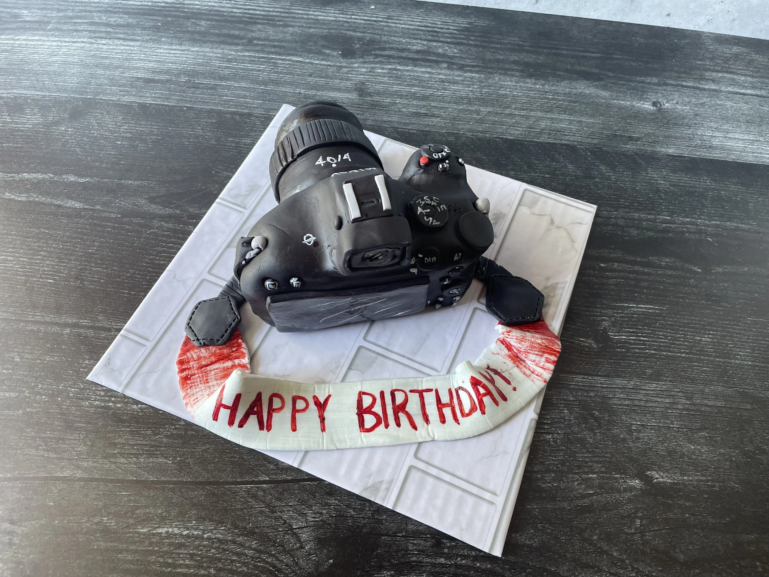 Camera-shaped birthday cake with a "Happy Birthday" message on a fondant banner, placed on a white tile mat on a dark wooden table.