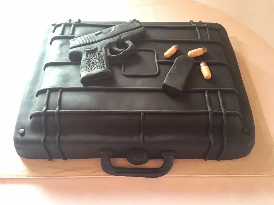 Cake shaped like a black suitcase with a gun, bullets, and gun accessories on top.