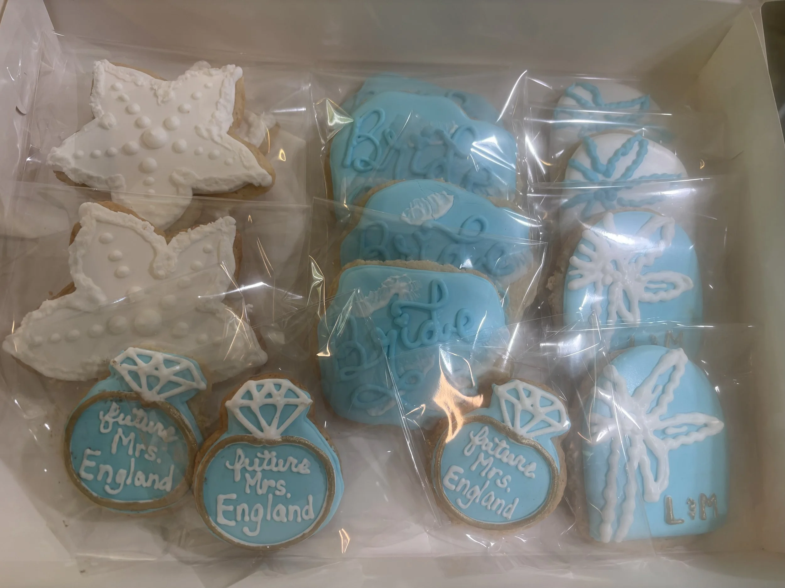 A box of decorated cookies with white, blue, and gold icing. The cookies feature starfish, hearts with the phrase 'future Mrs. England,' and other beach-themed designs.