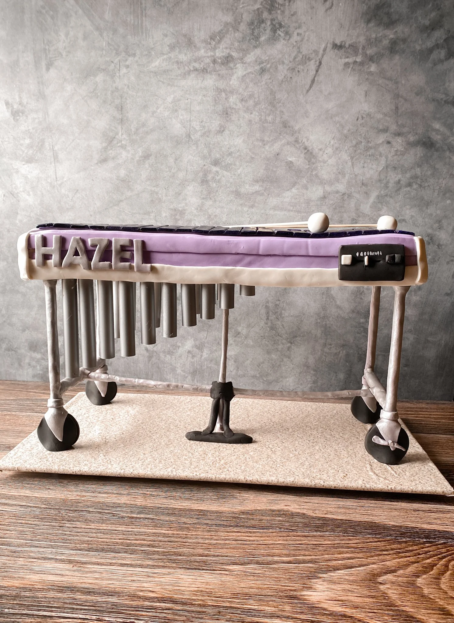 A cake shaped like a xylophone with the name "Hazel" written on the side, placed on a wooden table against a gray wall.