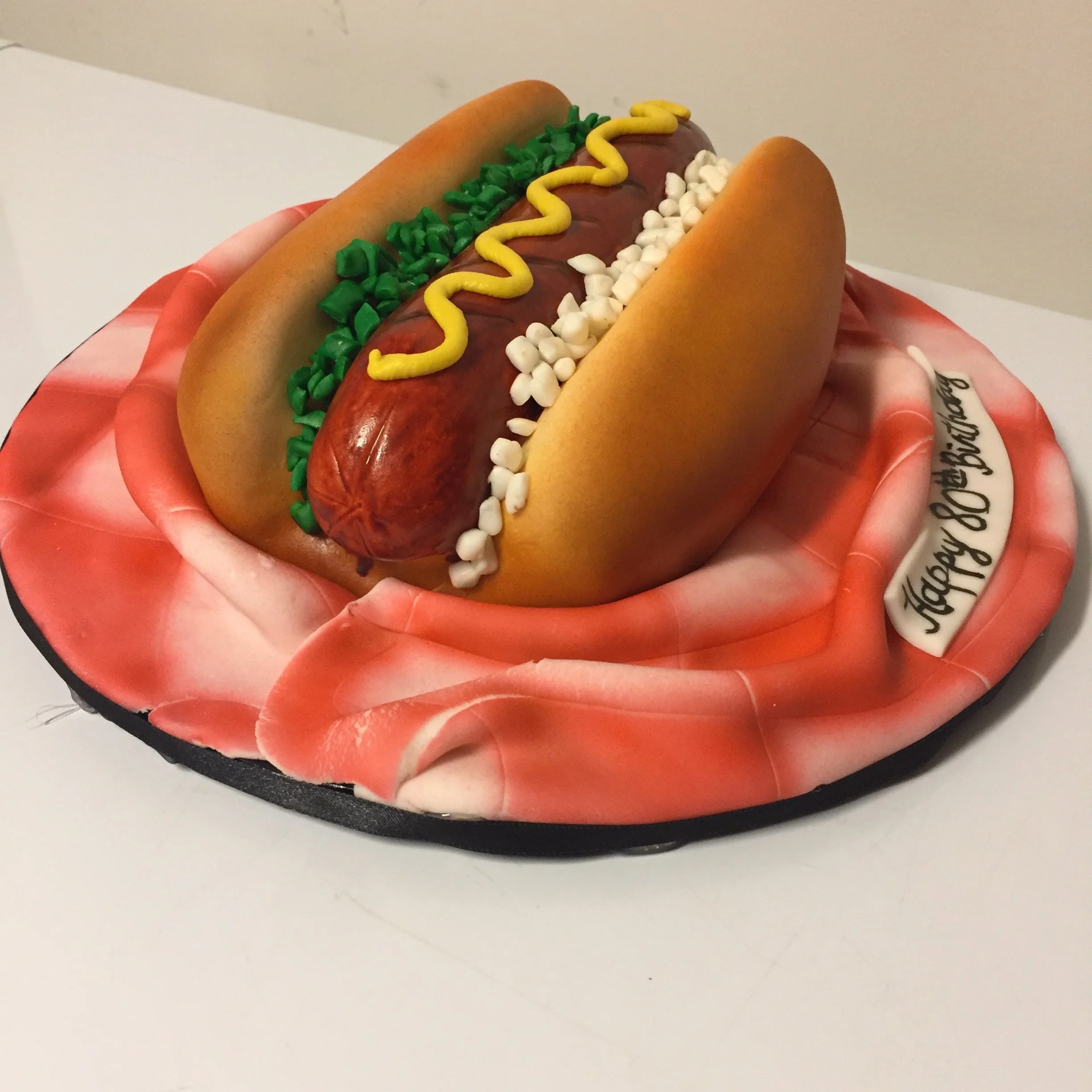 A cake designed to look like a hot dog in a bun, with mustard, relish, chopped onions, with a "Happy Birthday" message.