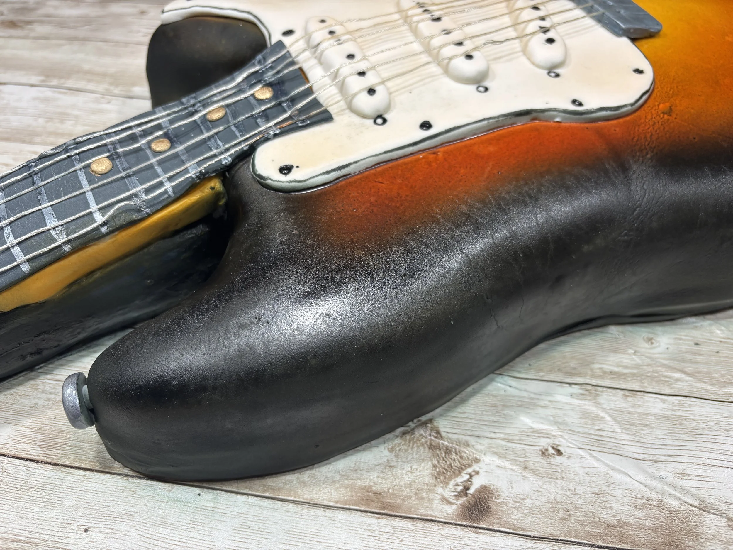 Close-up view of a realistic Fender Stratocaster electric guitar cake by Artful Cakery by Julie, showcasing hand-painted sunburst colors, edible knobs, strings, and textured details that mimic an authentic vintage guitar. Custom sculpted cake for wed