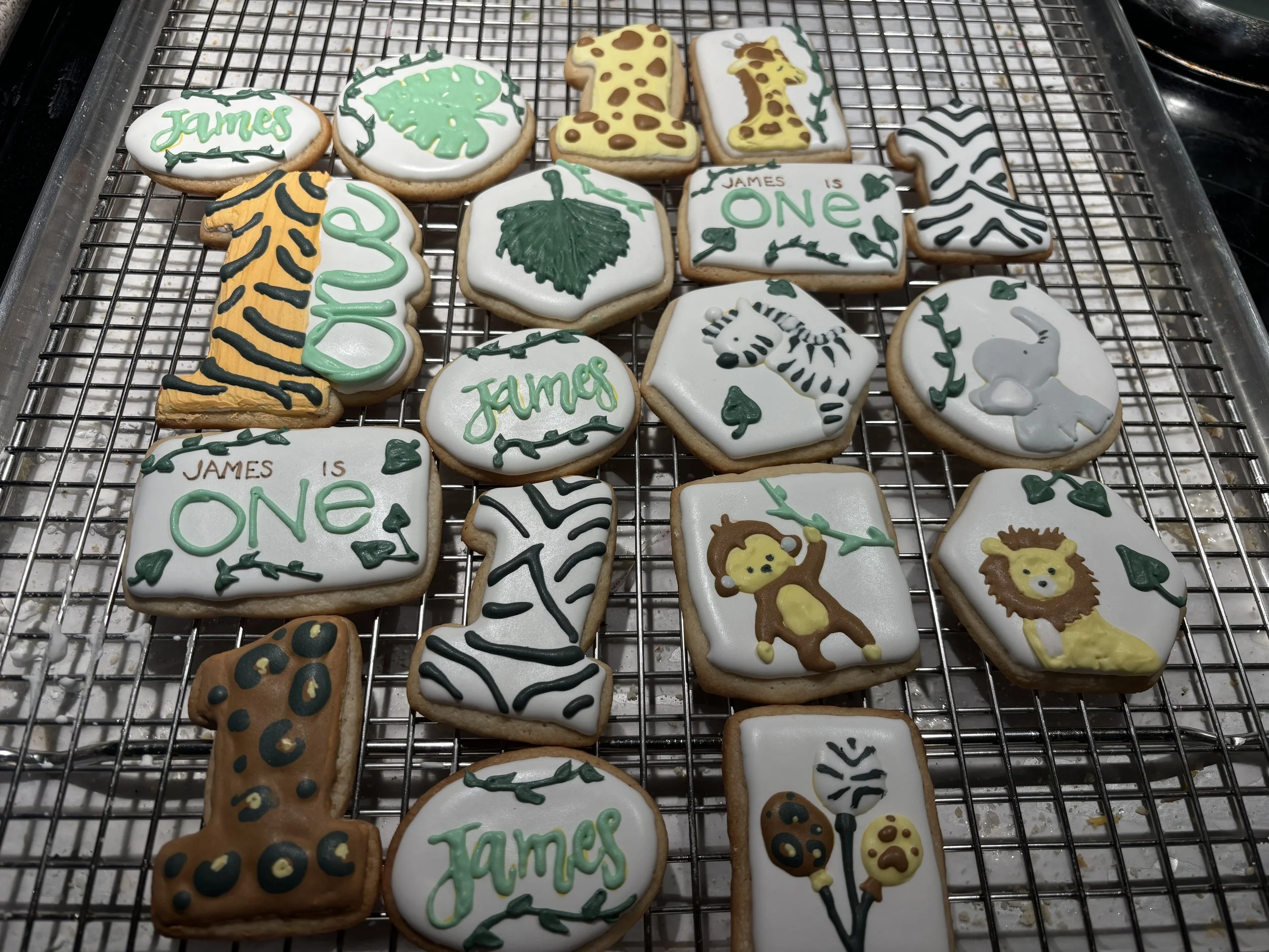 Assorted decorated cookies with jungle and zoo animal theme, including giraffe, zebra, elephant, lion, monkey, and leopard, some with text like 'James' and 'One', all on a metal cooling rack.