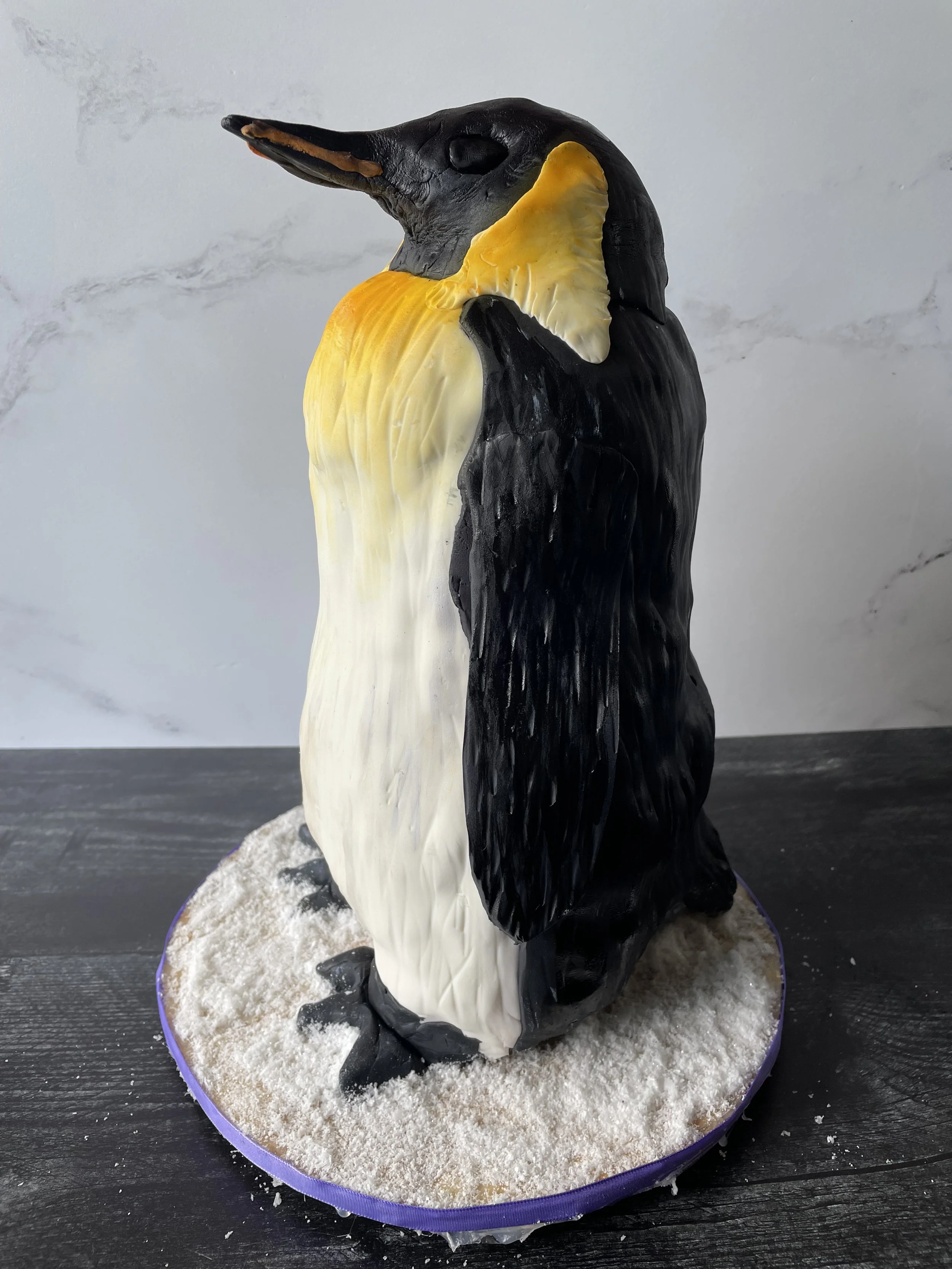 A cake decorated as a penguin with black, white, and yellow icing, standing on a snowy-looking base.
