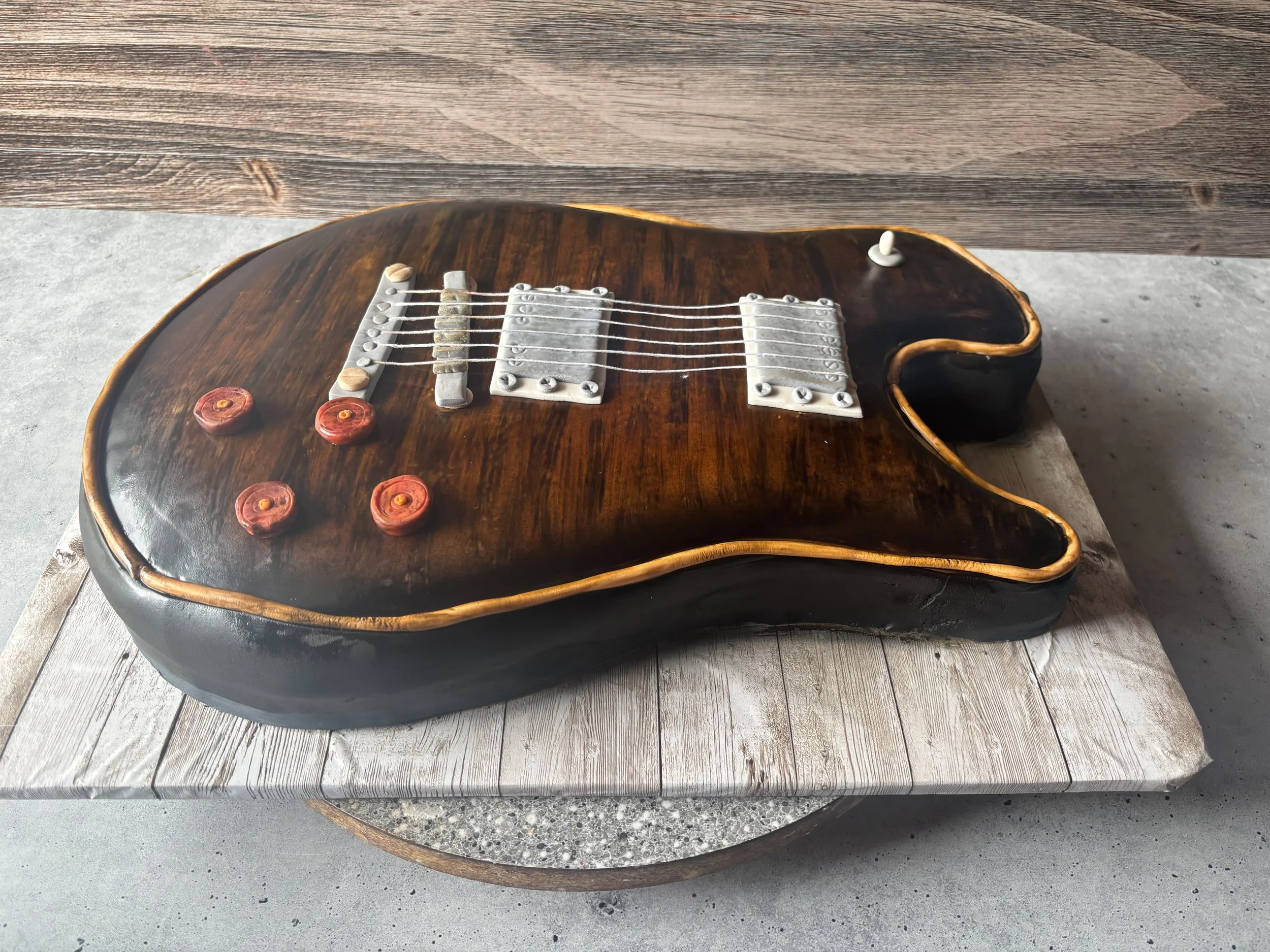 Cake shaped like a black electric guitar with brown wood-like details and red knobs, placed on a rustic wooden and concrete surface.