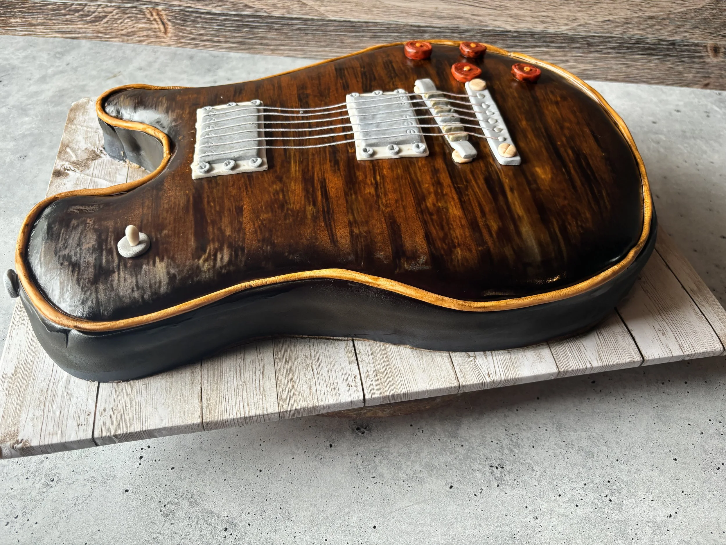A cake shaped like a vintage electric guitar with a brown wood grain design, white pickups, and red knobs, resting on a wooden surface.