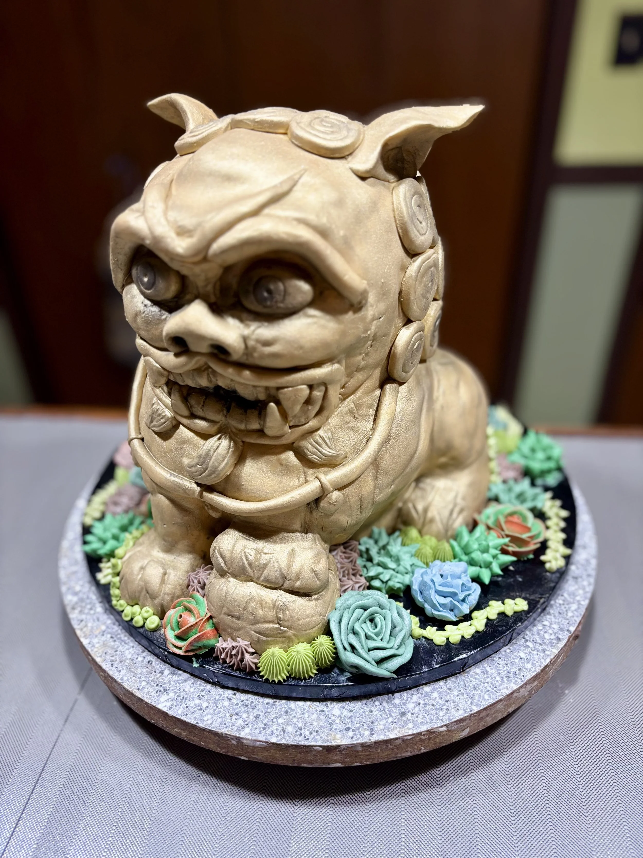 A detailed cake shaped like a Chinese dragon with multiple eyes, sharp teeth, and a fierce expression, decorated with colorful flower and foliage frosting around the base.