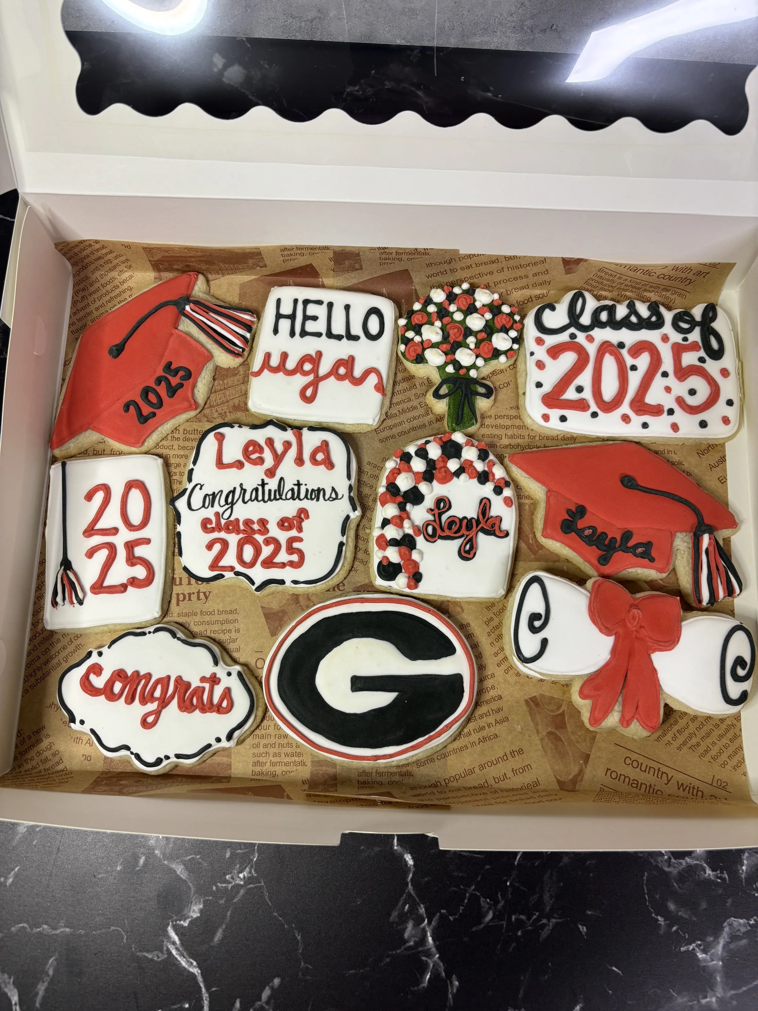 Box of decorated cookies celebrating graduation and a new school year, featuring caps, diplomas, bouquets, a letter G, and congratulatory messages.