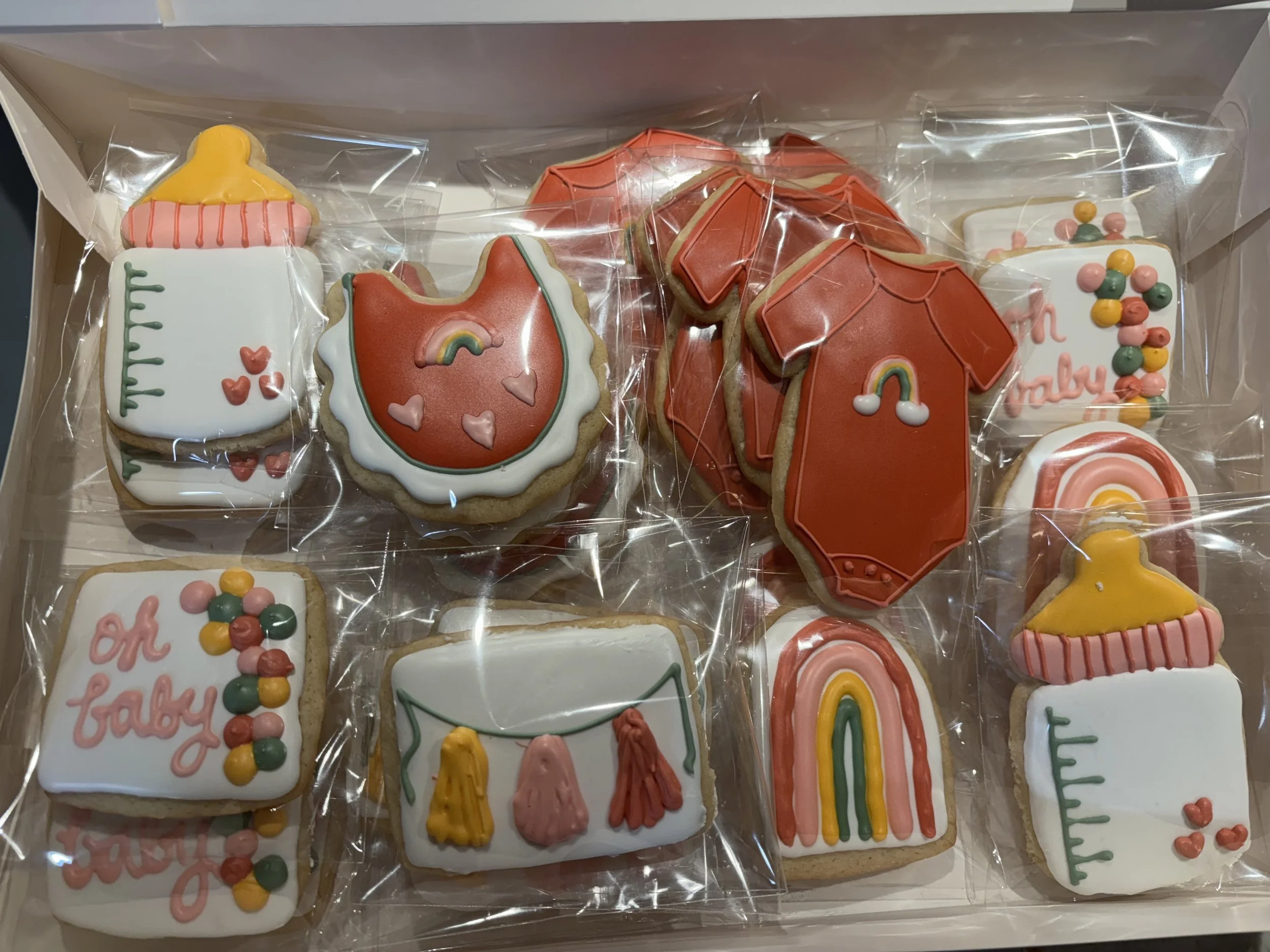 Assorted baby-themed decorated sugar cookies in a box, including baby bottles, onesies, rainbows decorated with pastel icing and fondant.