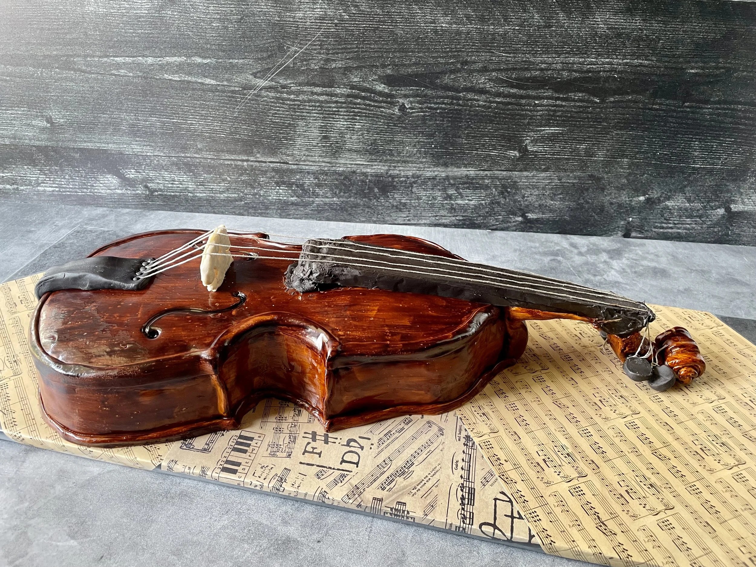 A violin-shaped cake with a glossy brown finish, decorated with fondant details representing strings and tuning pegs, placed on sheet music-themed paper on a gray surface.