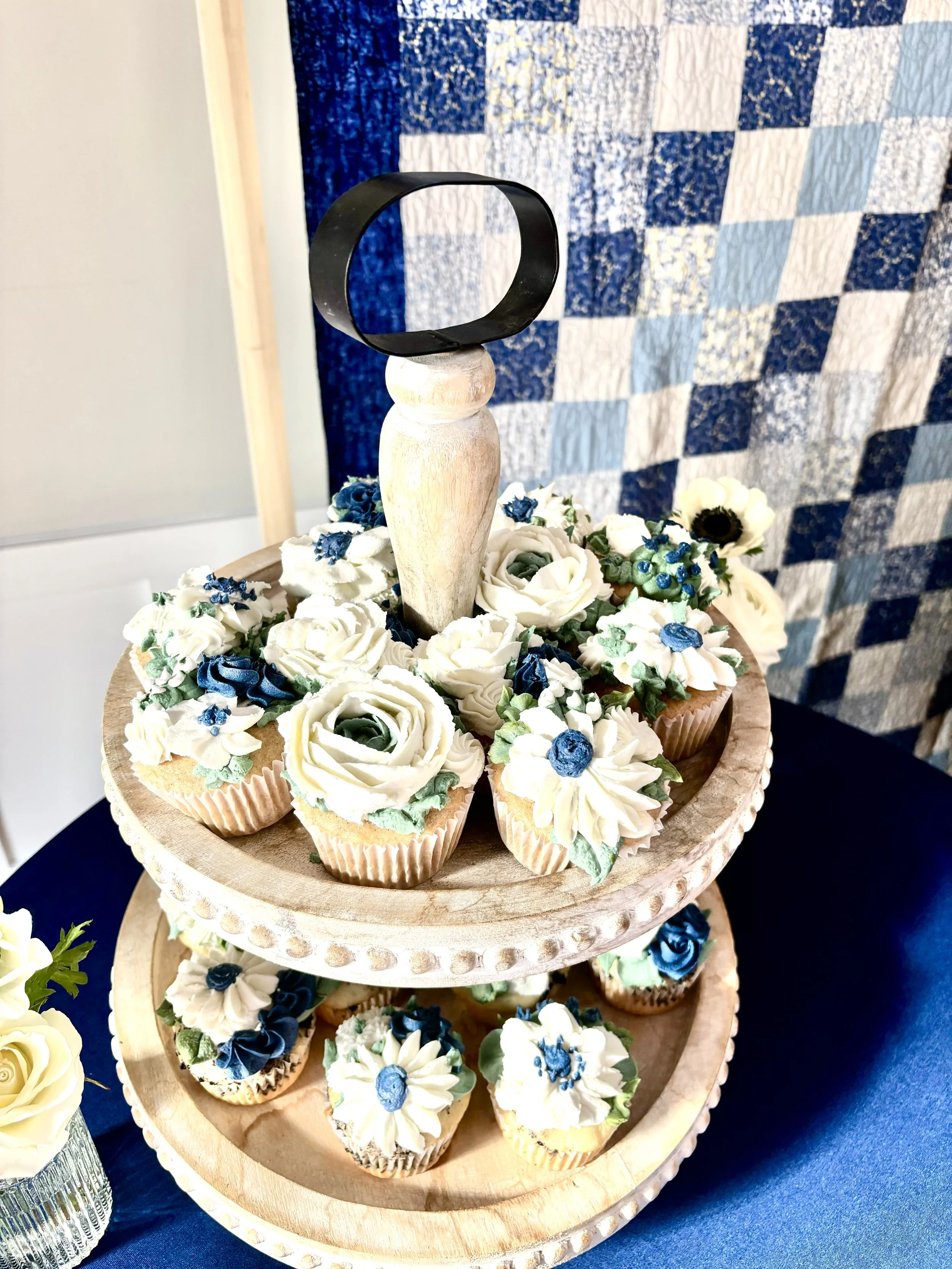 Wedding cupcakes decorated with blue and ivory buttercream roses, daisies, and greenery on a rustic wooden display at Juliette Chapel.