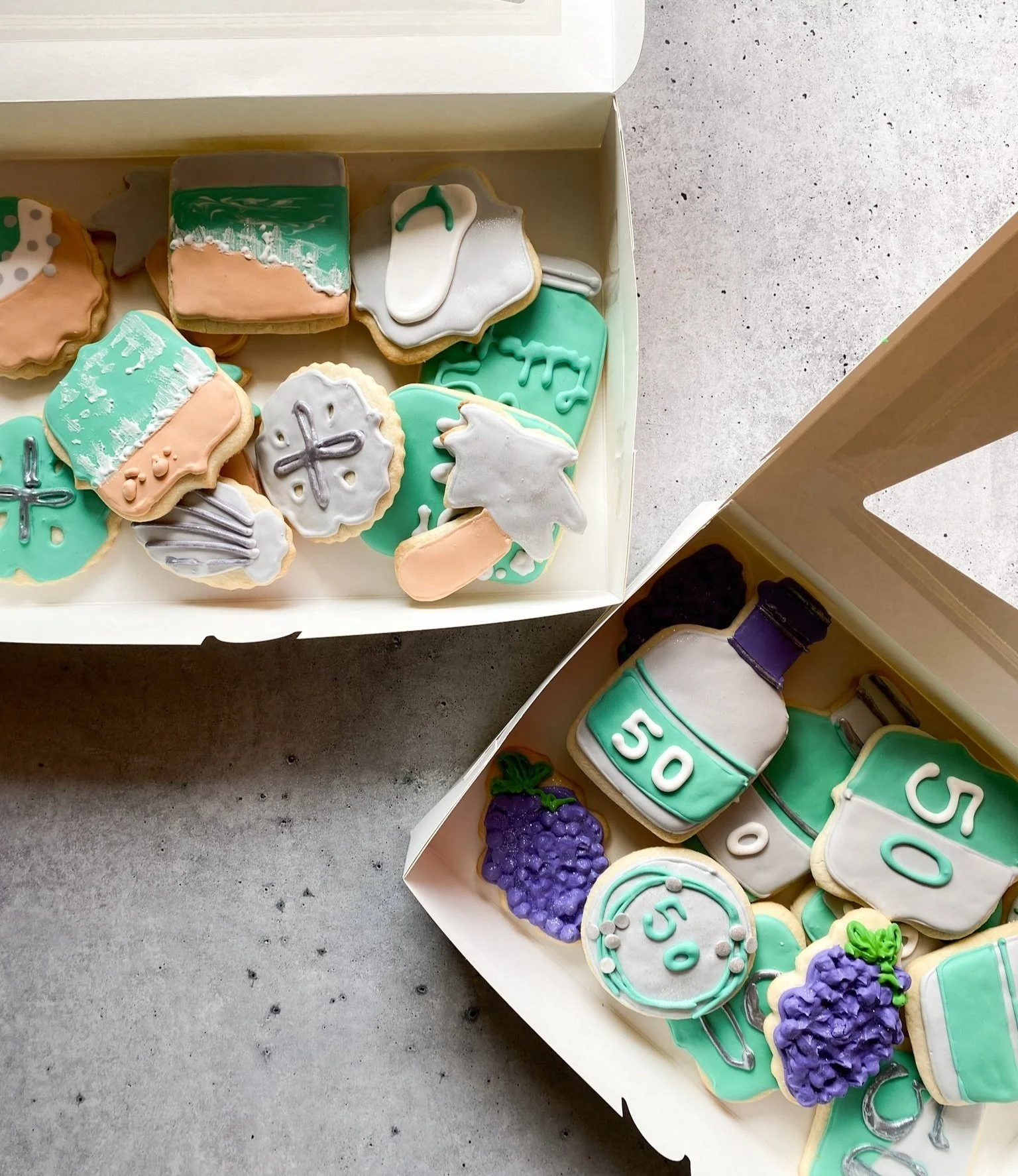 Two open boxes of decorated cookies, including cookies shaped like wine bottles, grapes, and various shapes with numbers and beverage-related decorations, on a concrete surface.