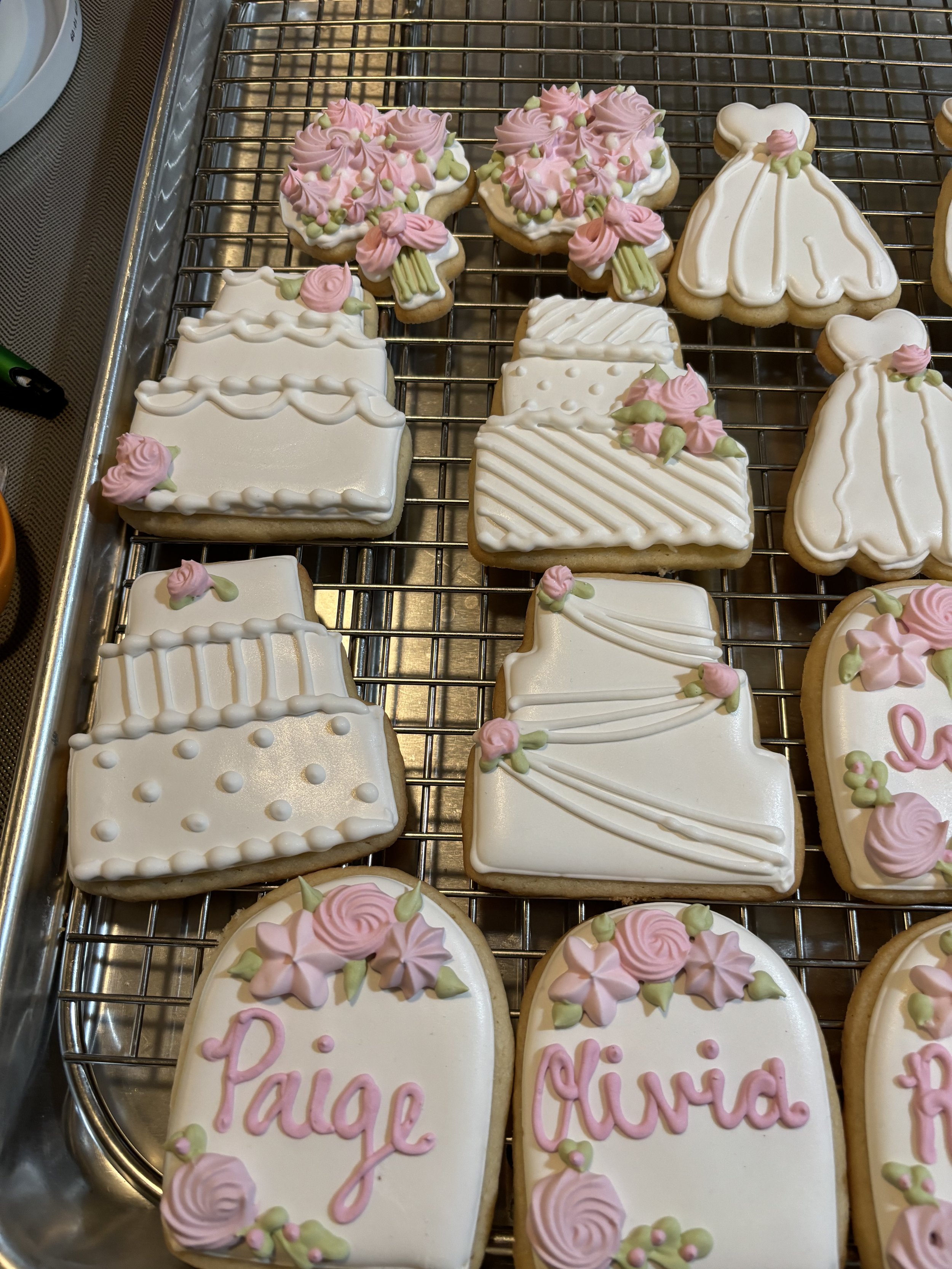 Decorated cookies in various shapes with white icing and pink and green floral decorations, some with names written on them in pink icing.