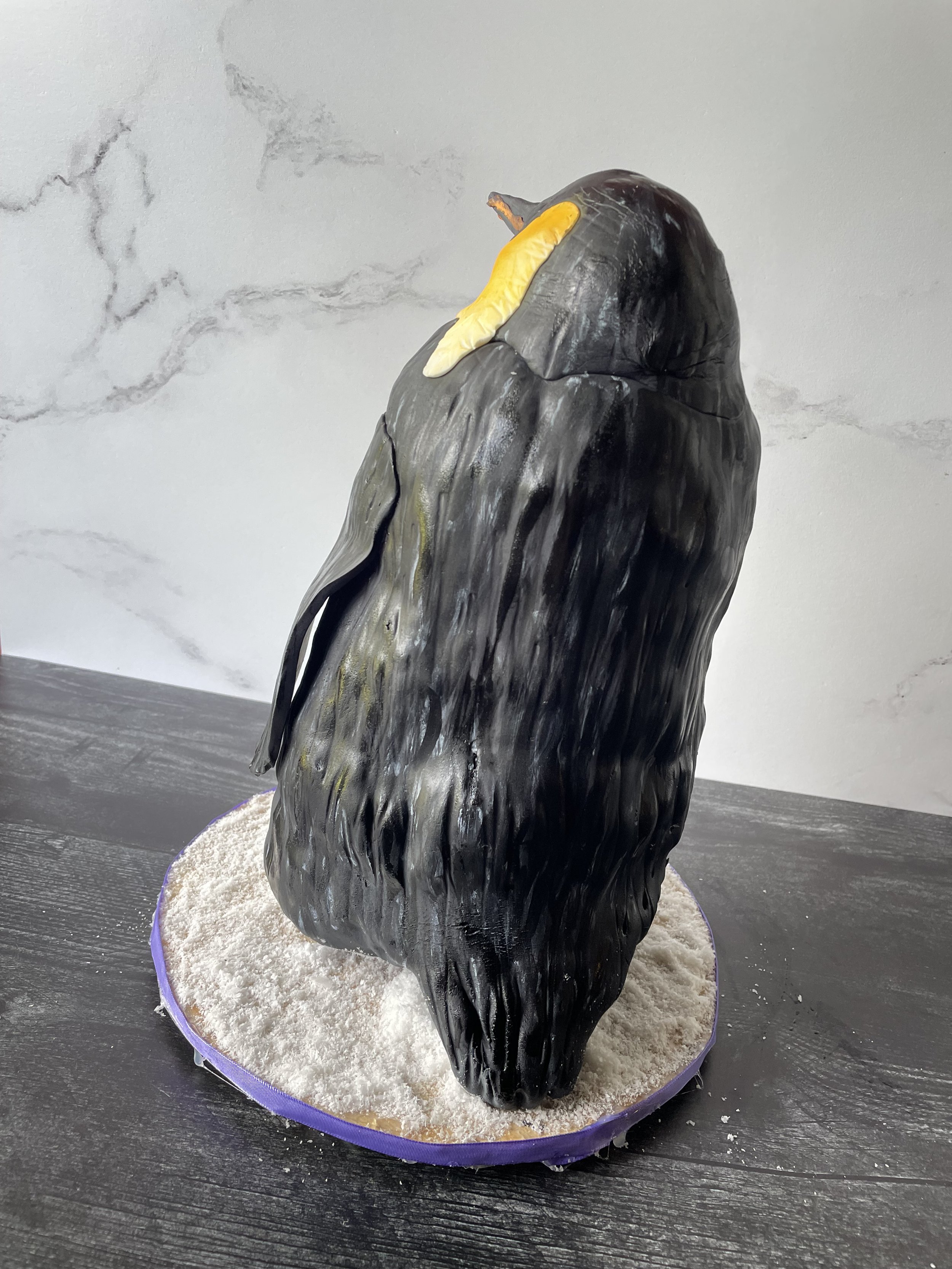 A cake designed to look like a penguin with yellow and orange accents on the beak, placed on a round purple and gold cake board with white powdery decoration resembling snow around it, on a dark wooden surface.