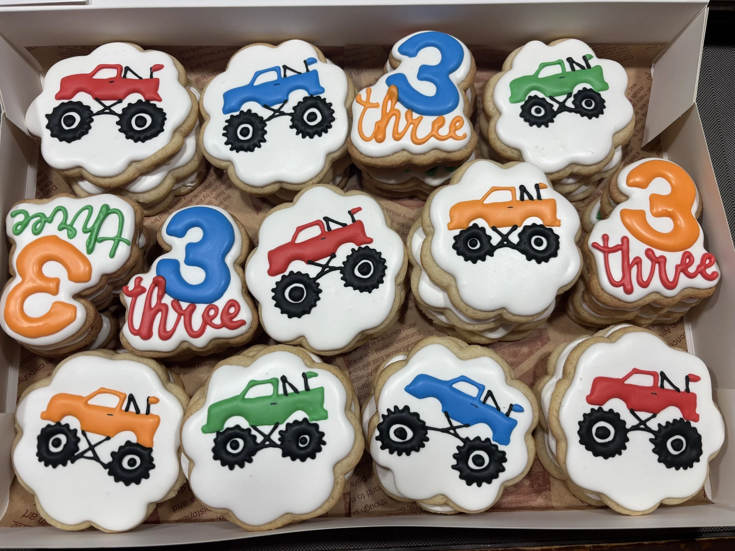 Collection of decorated cookies featuring colorful monster trucks, with the number three and the word 'three' in red, orange, green, blue, and black icing.