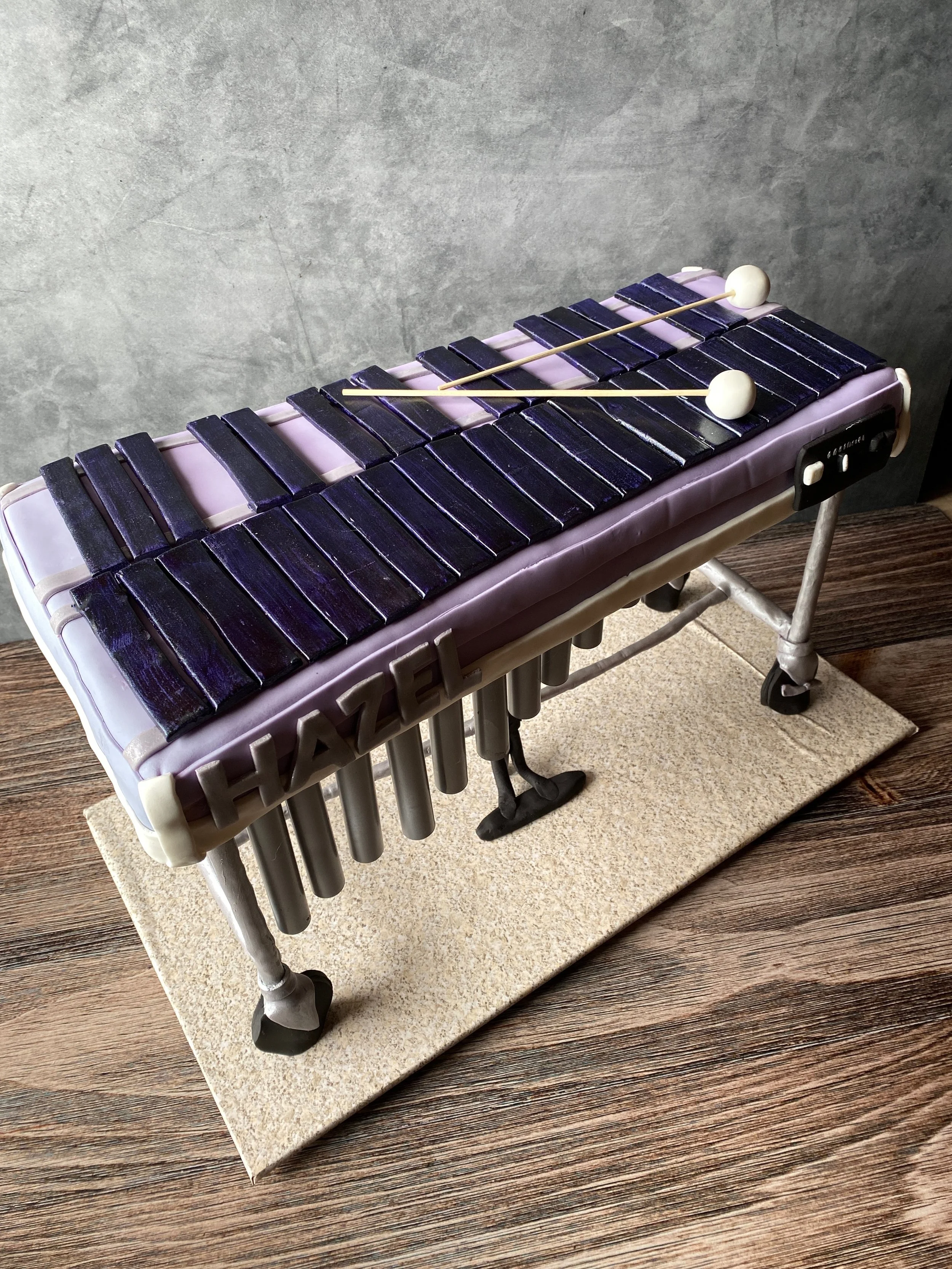 A cake designed to look like a xylophone, with black and purple bars, white mallets on top, and the word 'HAZEL' spelled out on the side.