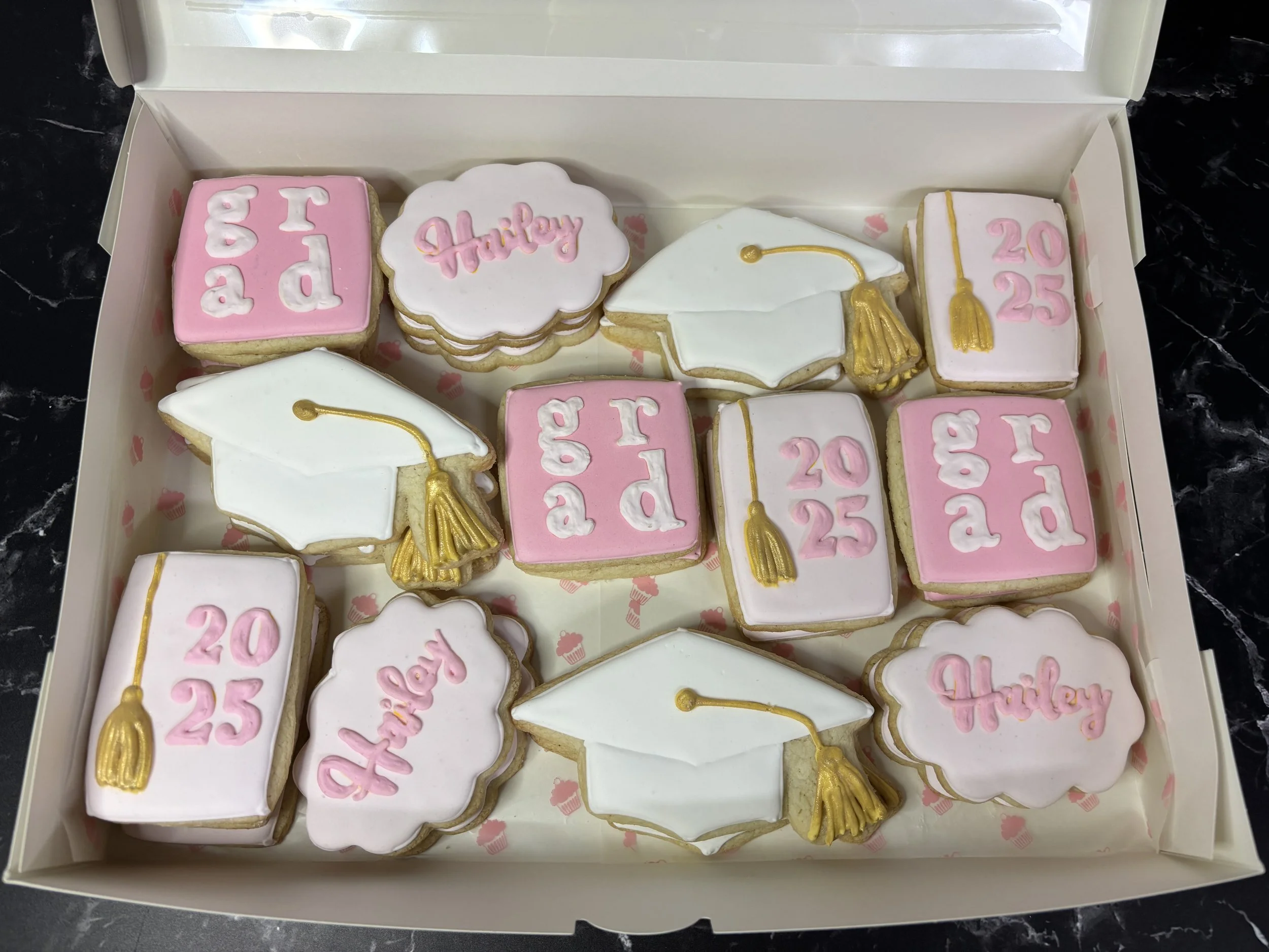 A box of decorated cookies celebrating a graduation and 2025 graduation year, with some cookies shaped like graduation caps, some with the word 'Hailey, and others with 'grad' and the year.