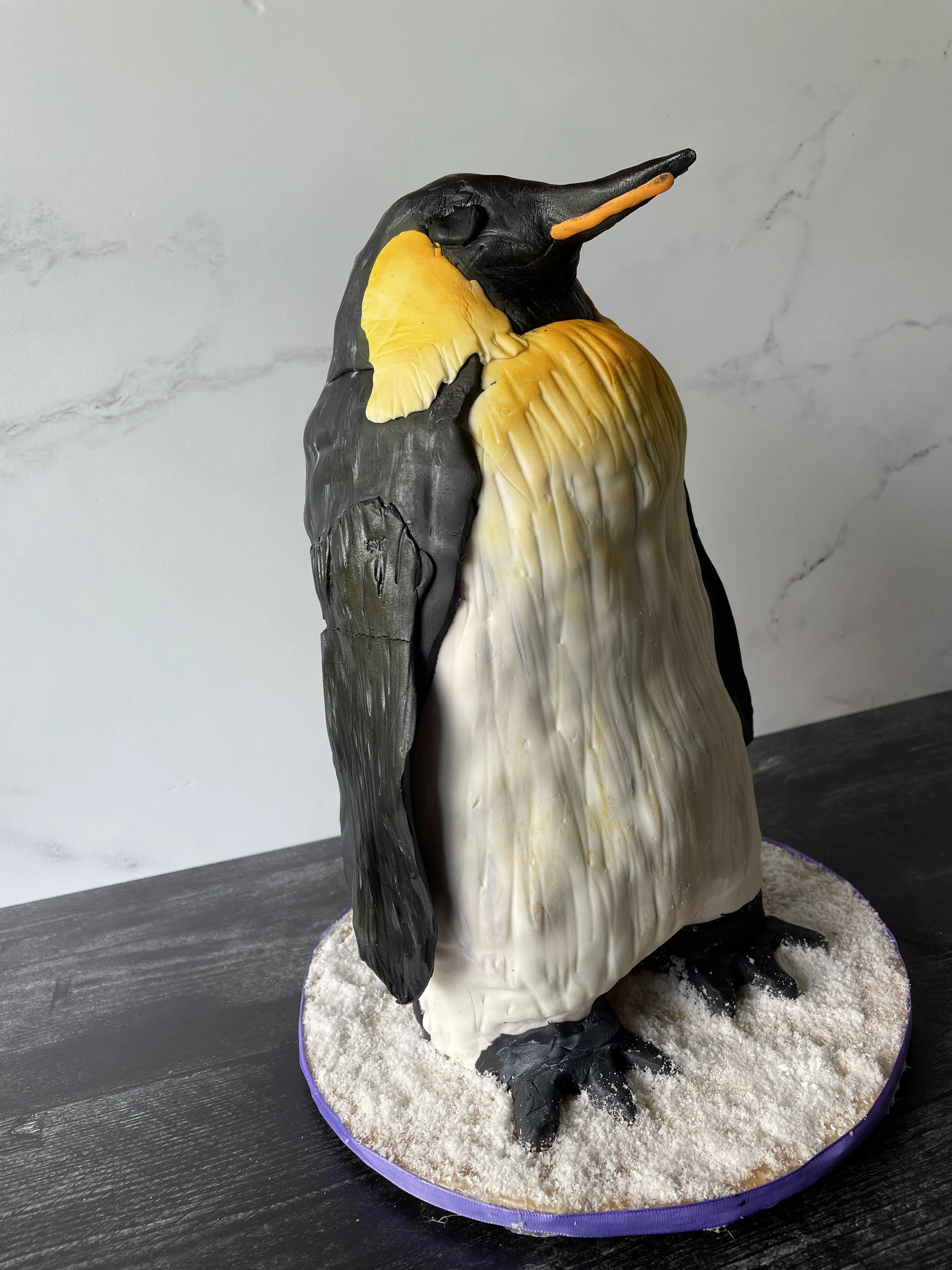 A penguin-shaped cake decorated with black, yellow, and white icing, standing on a round base covered to resemble snow.