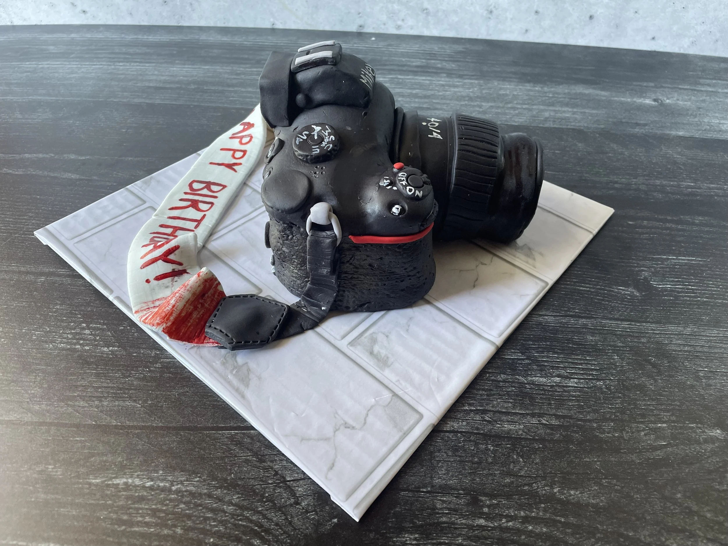 Cake designed to look like a camera with a white ribbon that says 'Happy Birthday' in red letters, placed on a white tiled cake board.