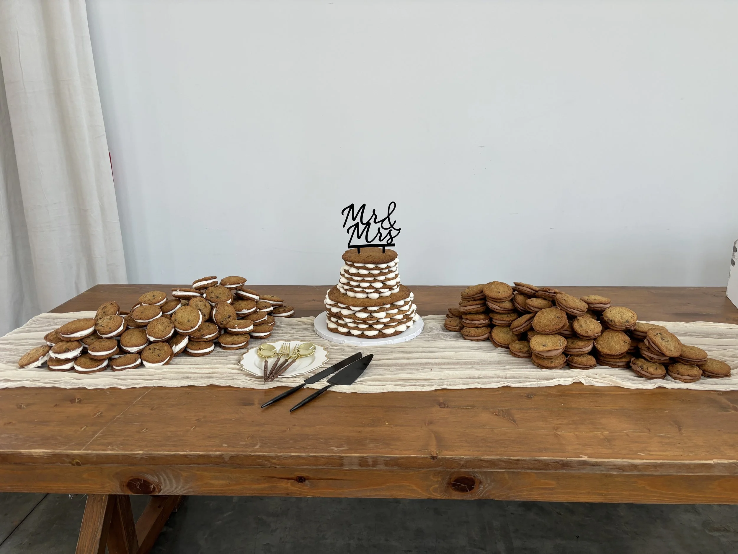A wedding dessert table with a stack of cookies in the center, topped with a topper reading "Mrs & Mrs." Flanking the stack are piles of cookies, with utensils and a serving tray in front of the cookies.