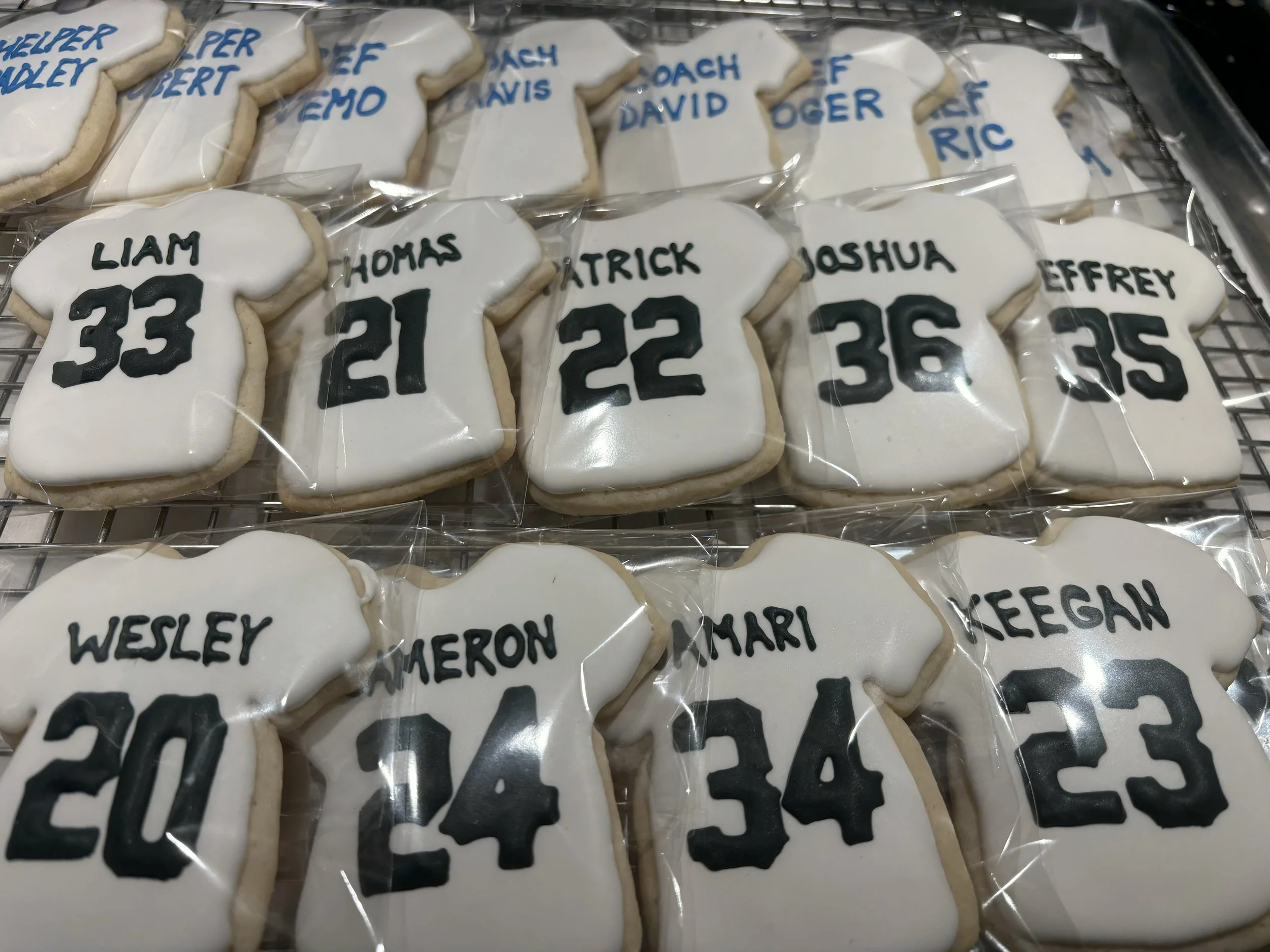 Sugar cookies decorated as sports jerseys with player names and numbers, individually wrapped in plastic. The cookies are displayed on a wire rack.