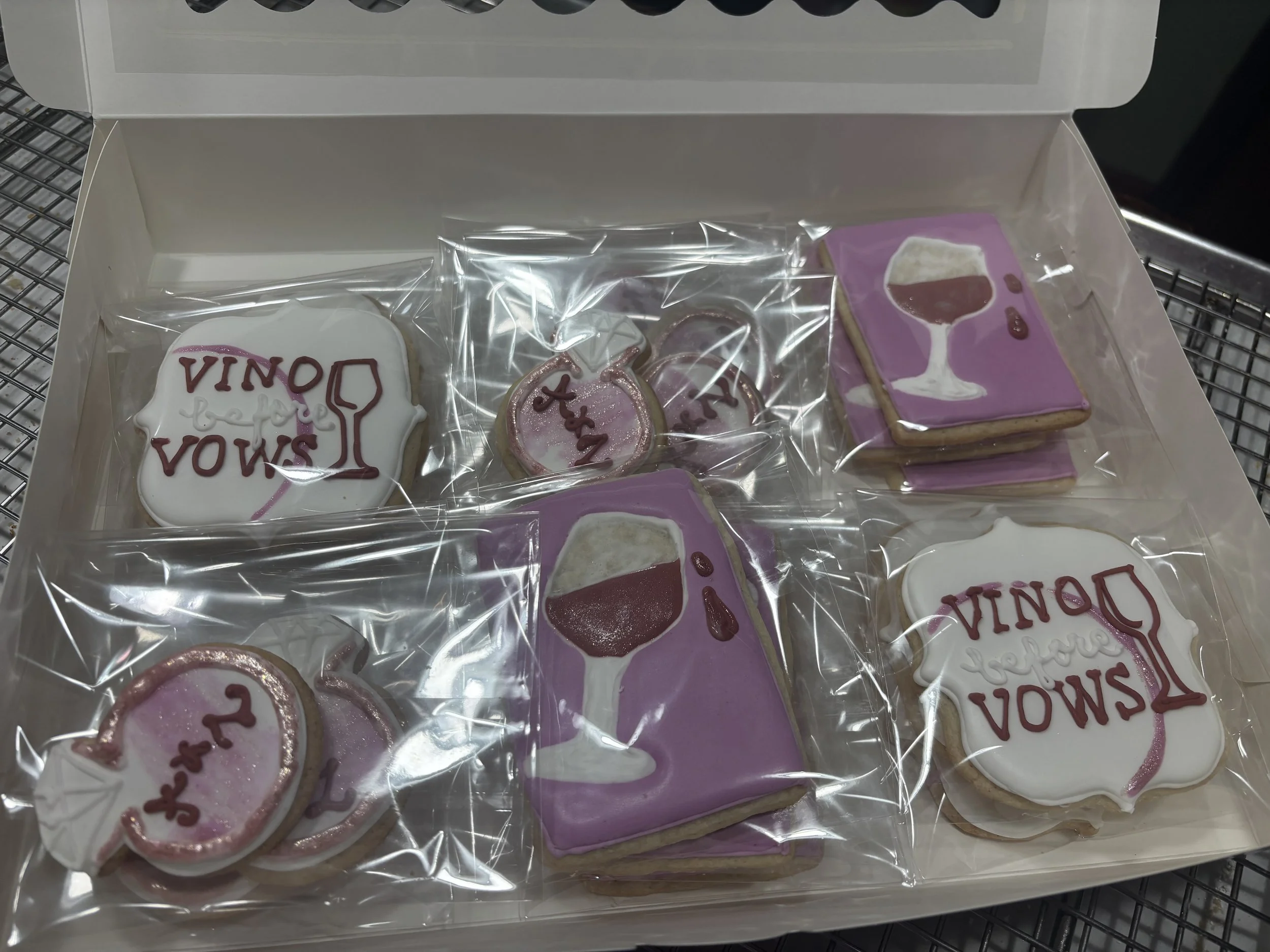Assorted decorated cookies in a box, featuring a wine glass, wine bottle, with purple and pink themes, wrapped individually in clear plastic.
