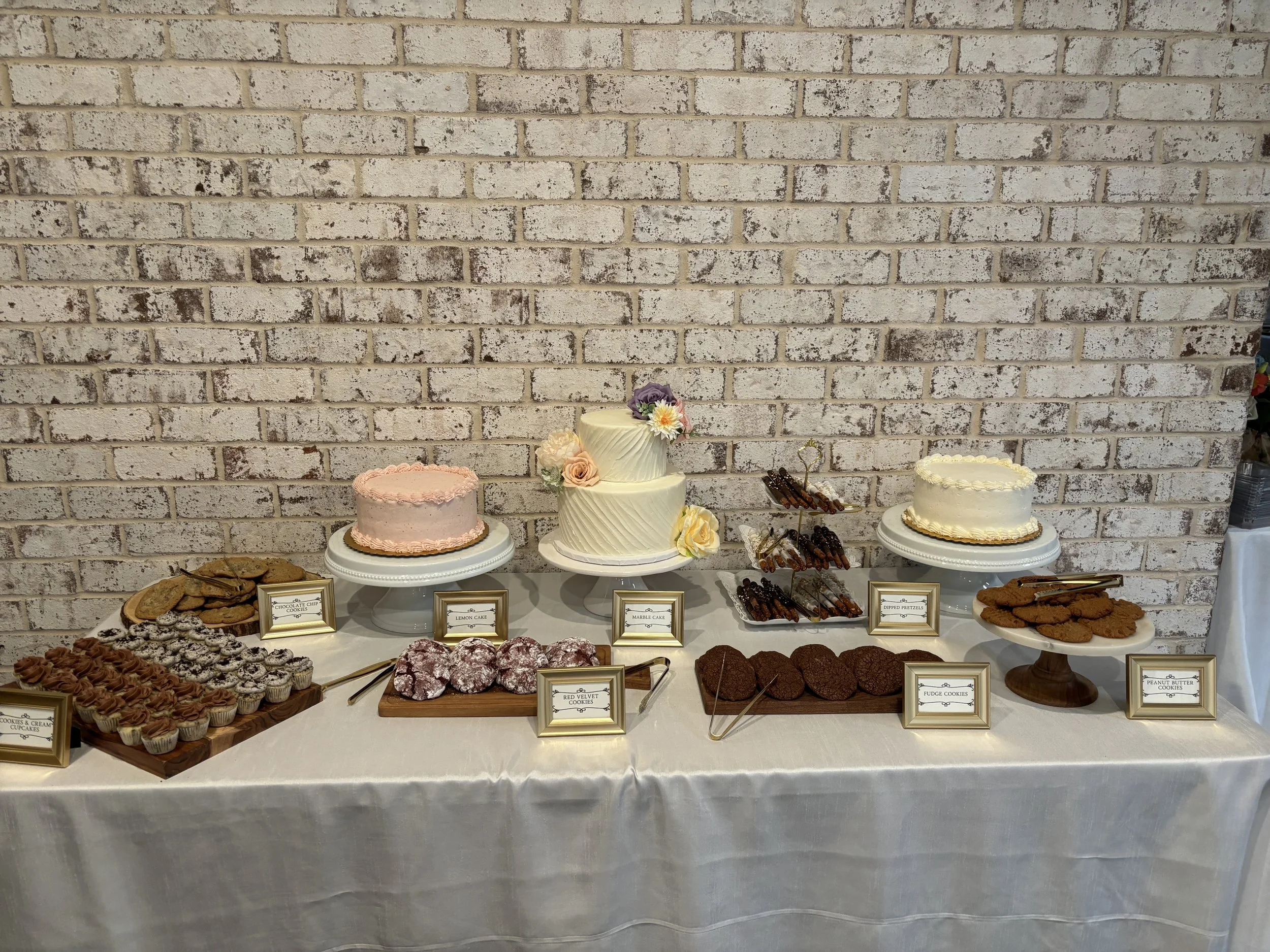 Dessert table with three cakes, cookies, brownies, and pretzels displayed against a brick wall