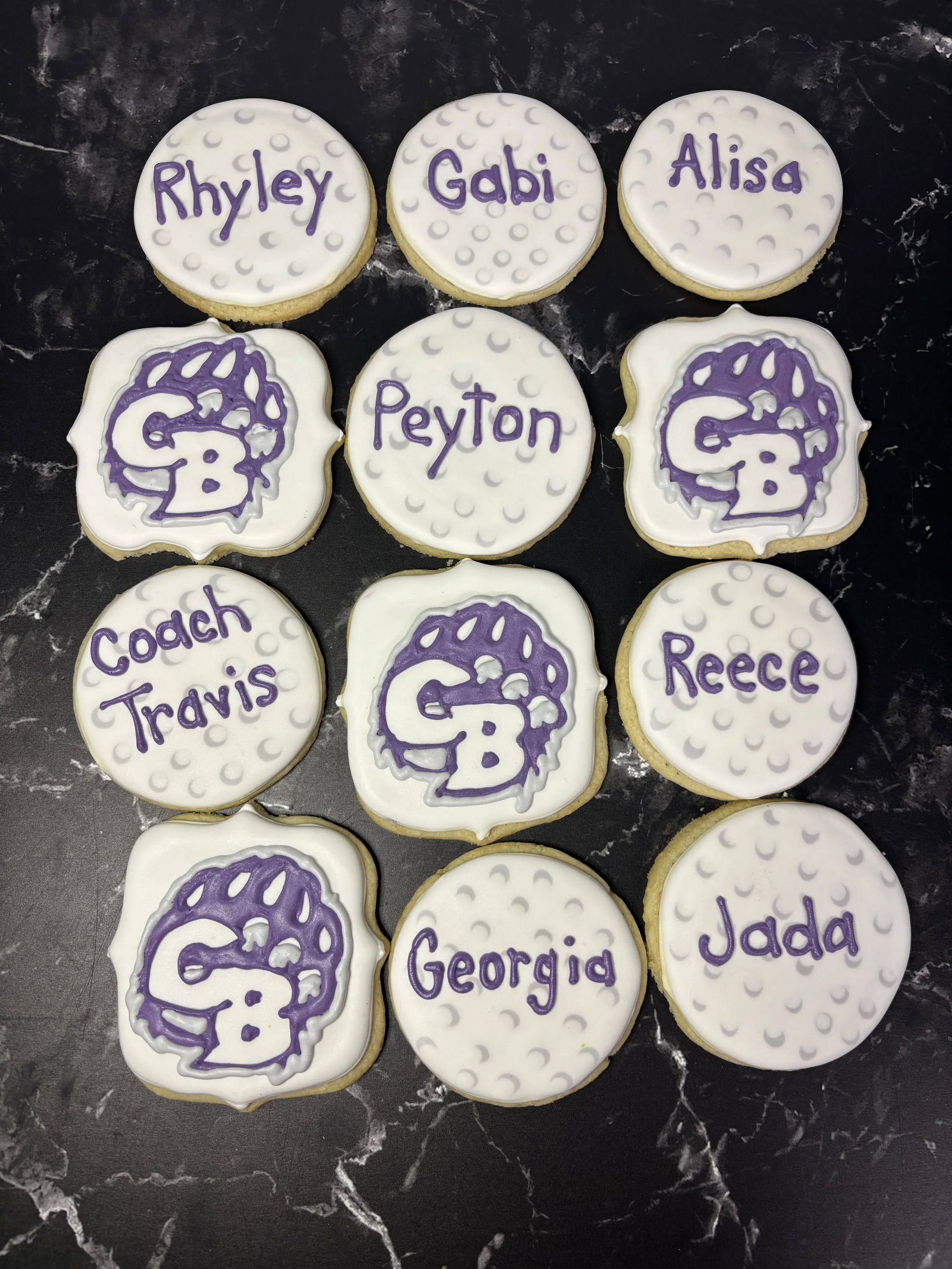 A collection of decorated golf ball cookies with names and a purple bearprint logo on white icing.