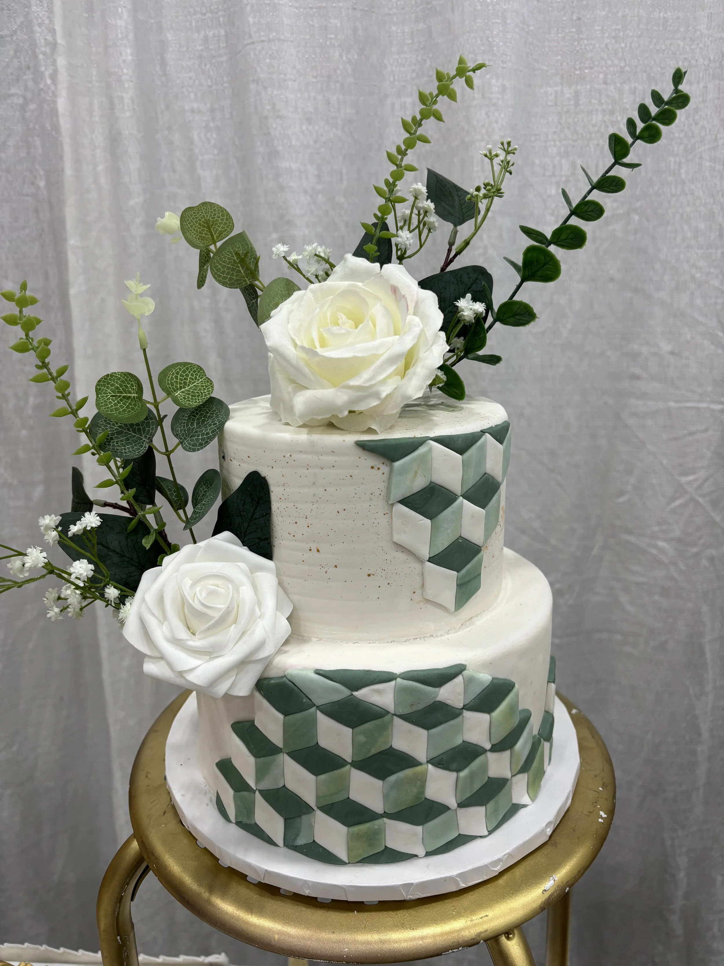 Two-tiered white wedding cake decorated with geometric green and white fondant patterns, adorned with white roses and greenery, placed on a small gold-colored stool.