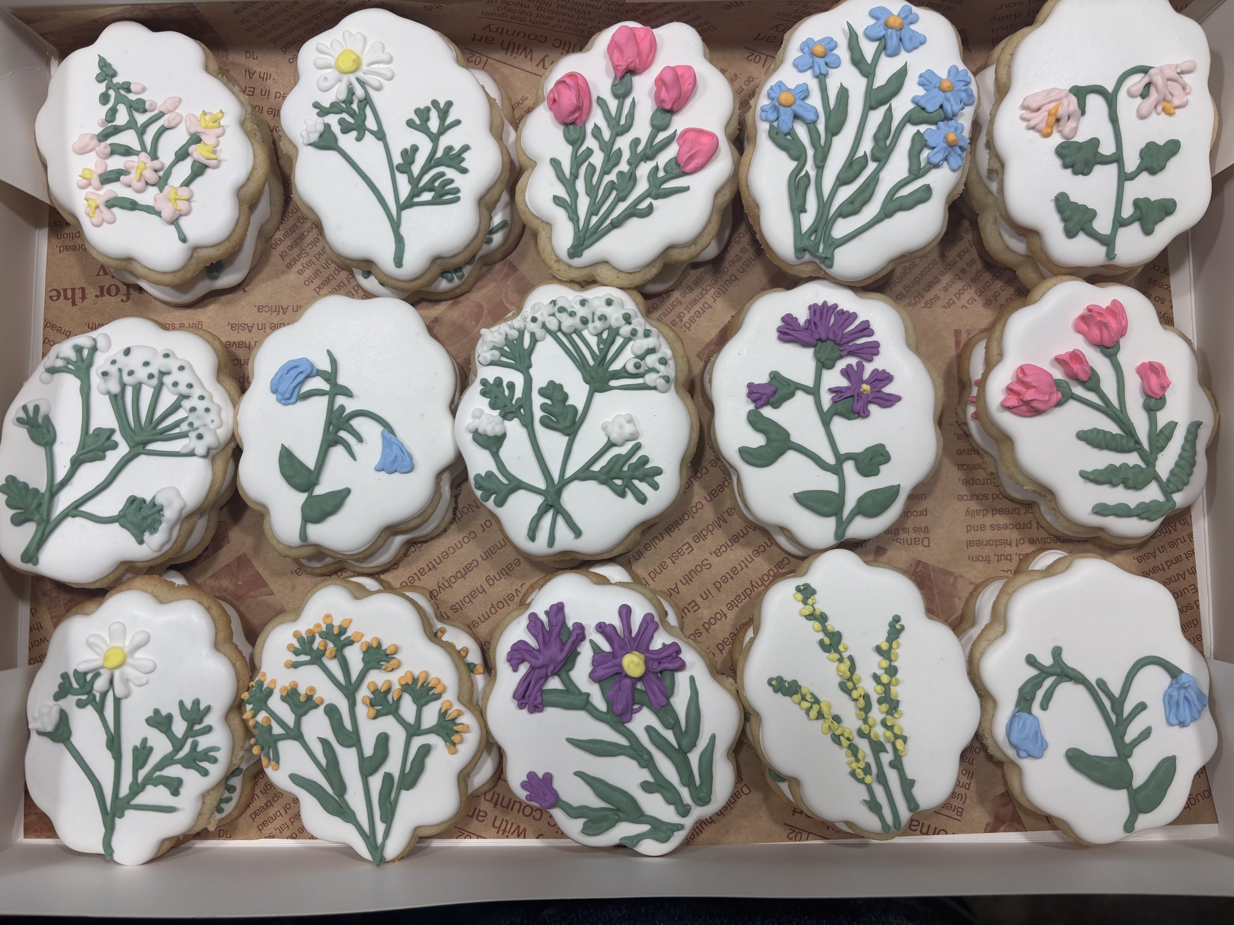 A box of decorated cookies with floral icing designs featuring various colorful flowers, including daisies, tulips, and others, on white icing
