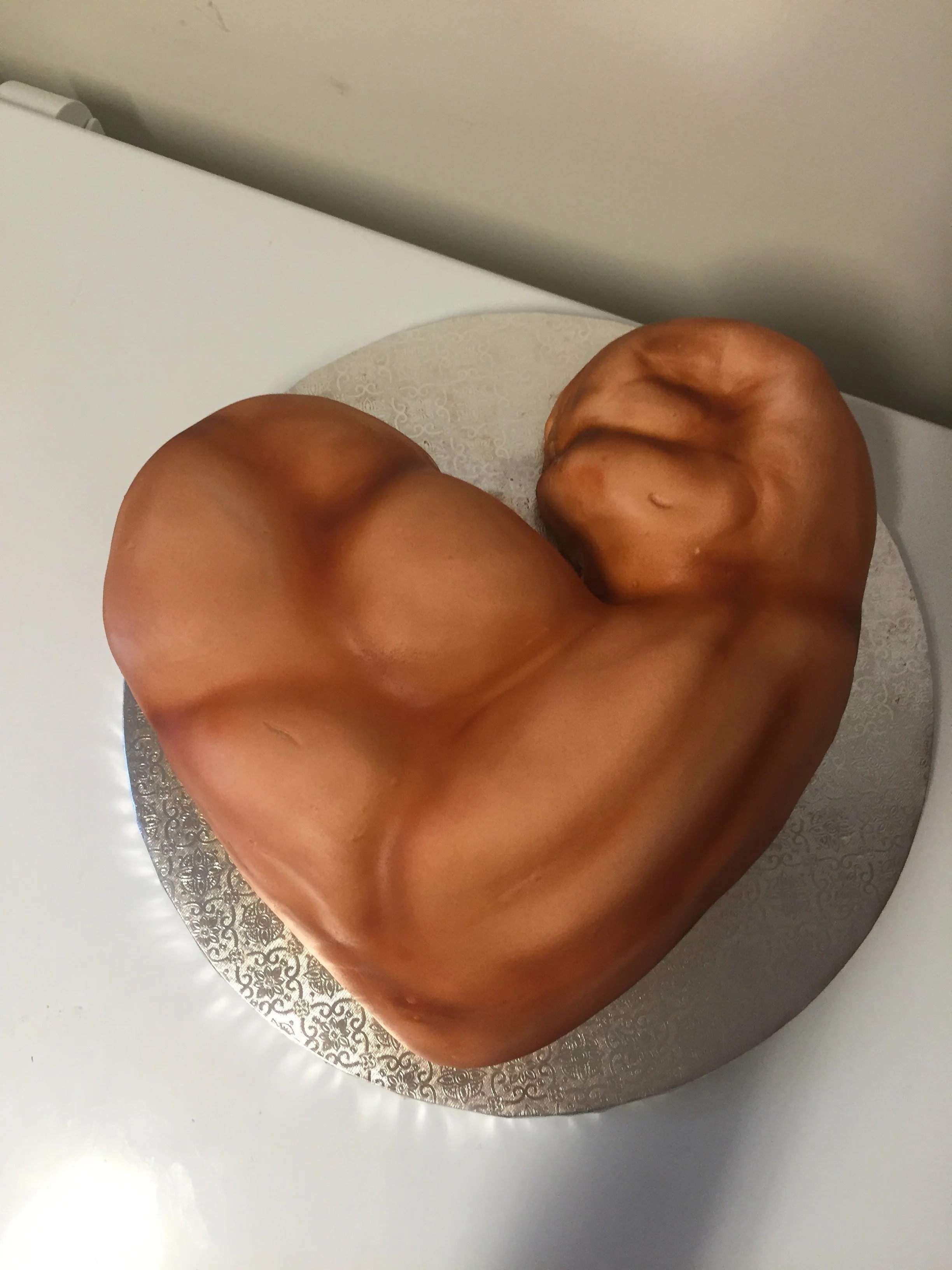 A cake shaped like a heart with a sculpted muscular arm on the side, placed on a round silver cake board.