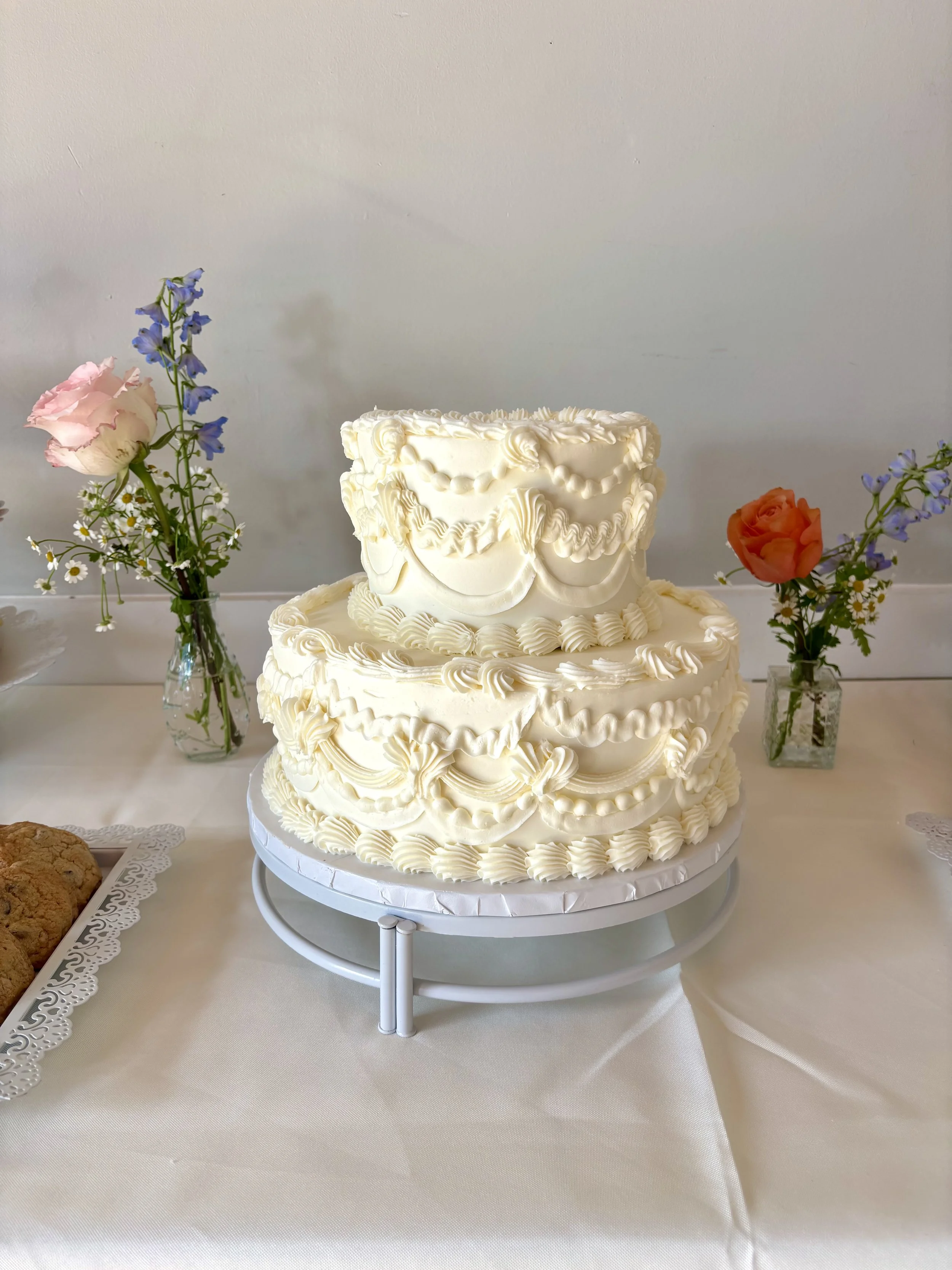 Ivory two-tier Lambeth-style buttercream wedding cake with ornate piped swags and ruffles, displayed with wedding cupcakes, cookies, and cake pops at Juliette Chapel.