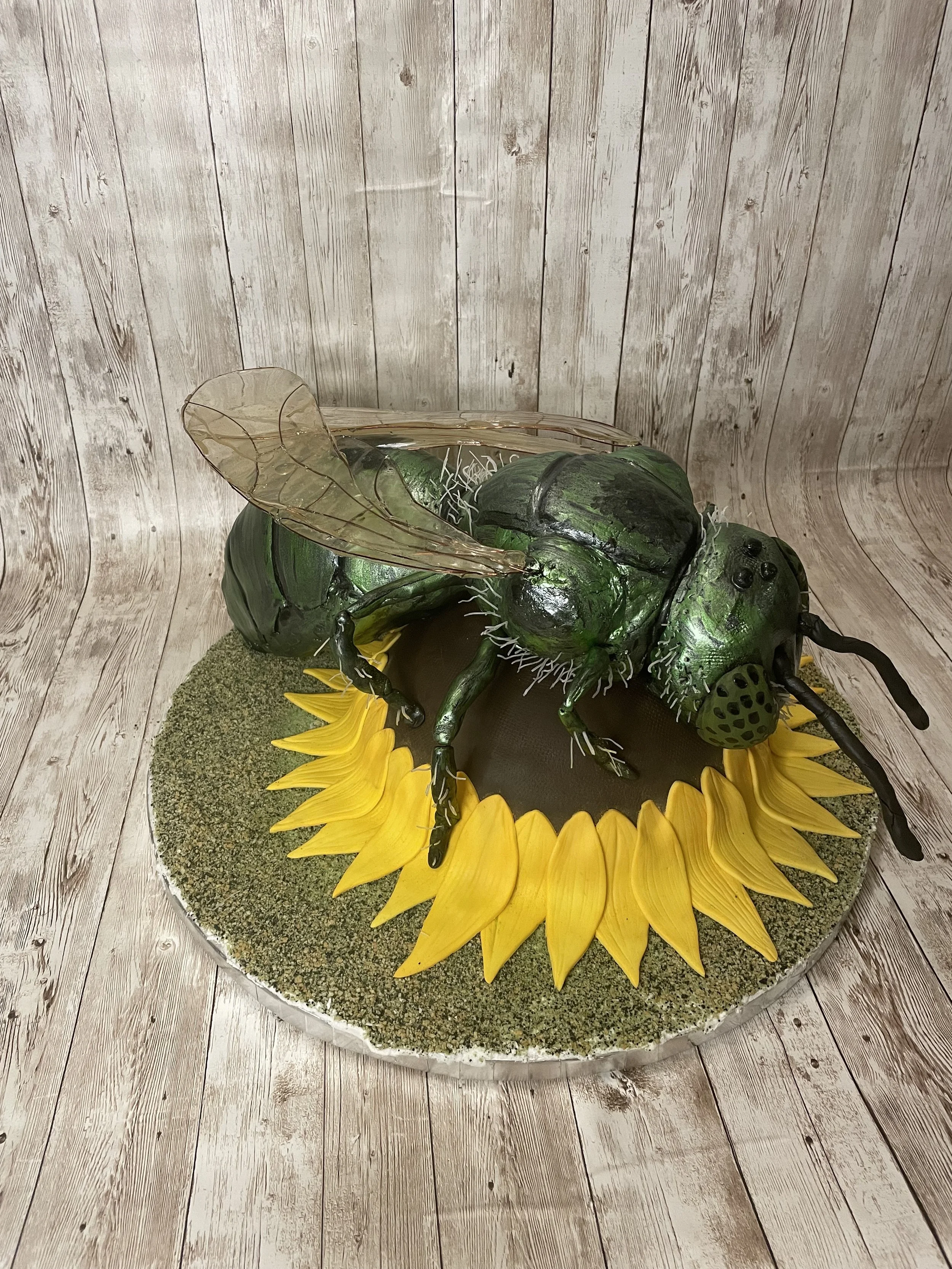 A cake of a large green metallic sweat bee with transparent wings, sitting on a sunflower cake. detailed features and a realistic appearance.