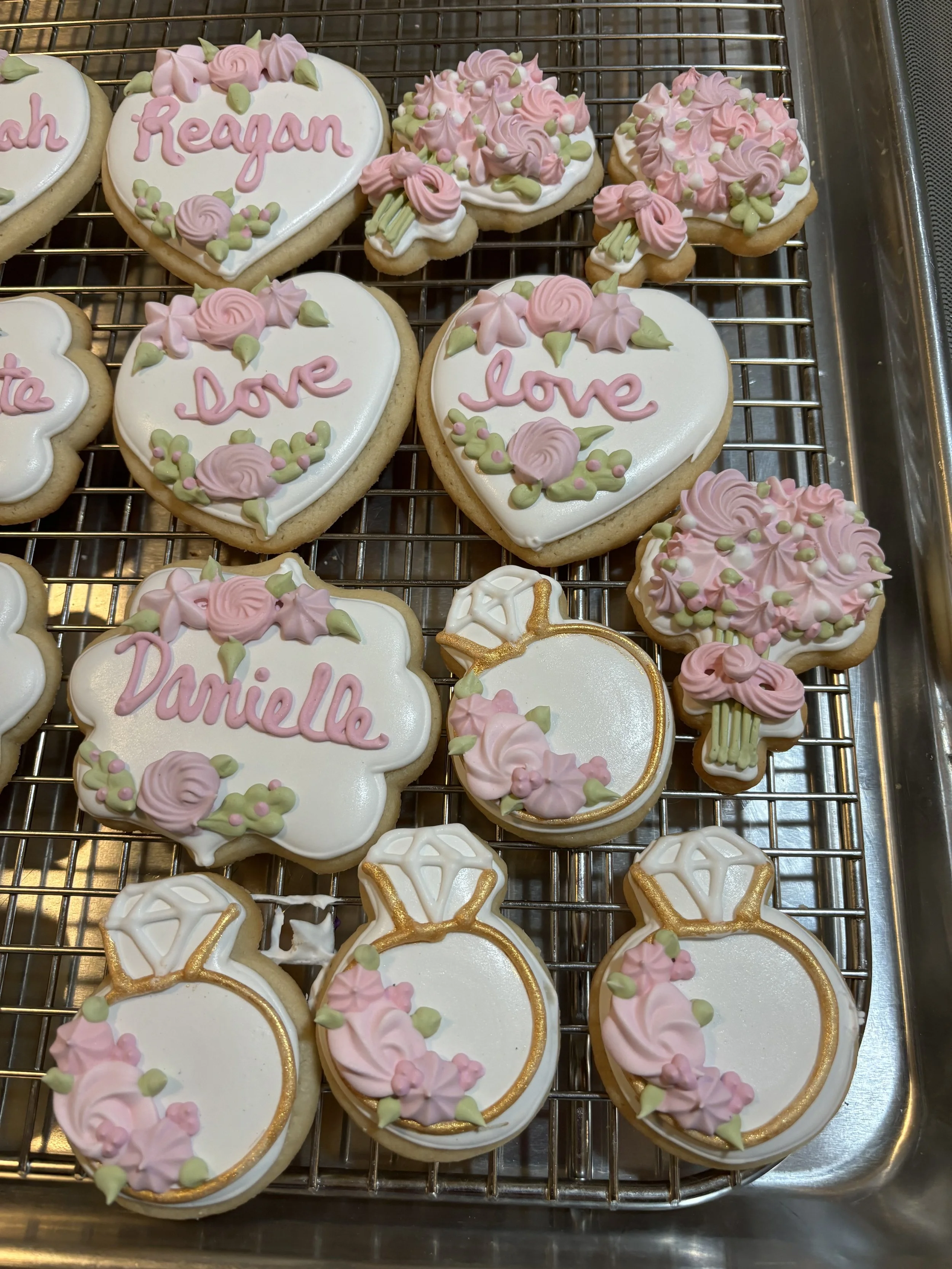 Assorted decorated cookies for a celebration, featuring hearts, flowers, and a diamond ring motif, with names and words like 'Reagan', 'love', and 'Danielle' written on some cookies.