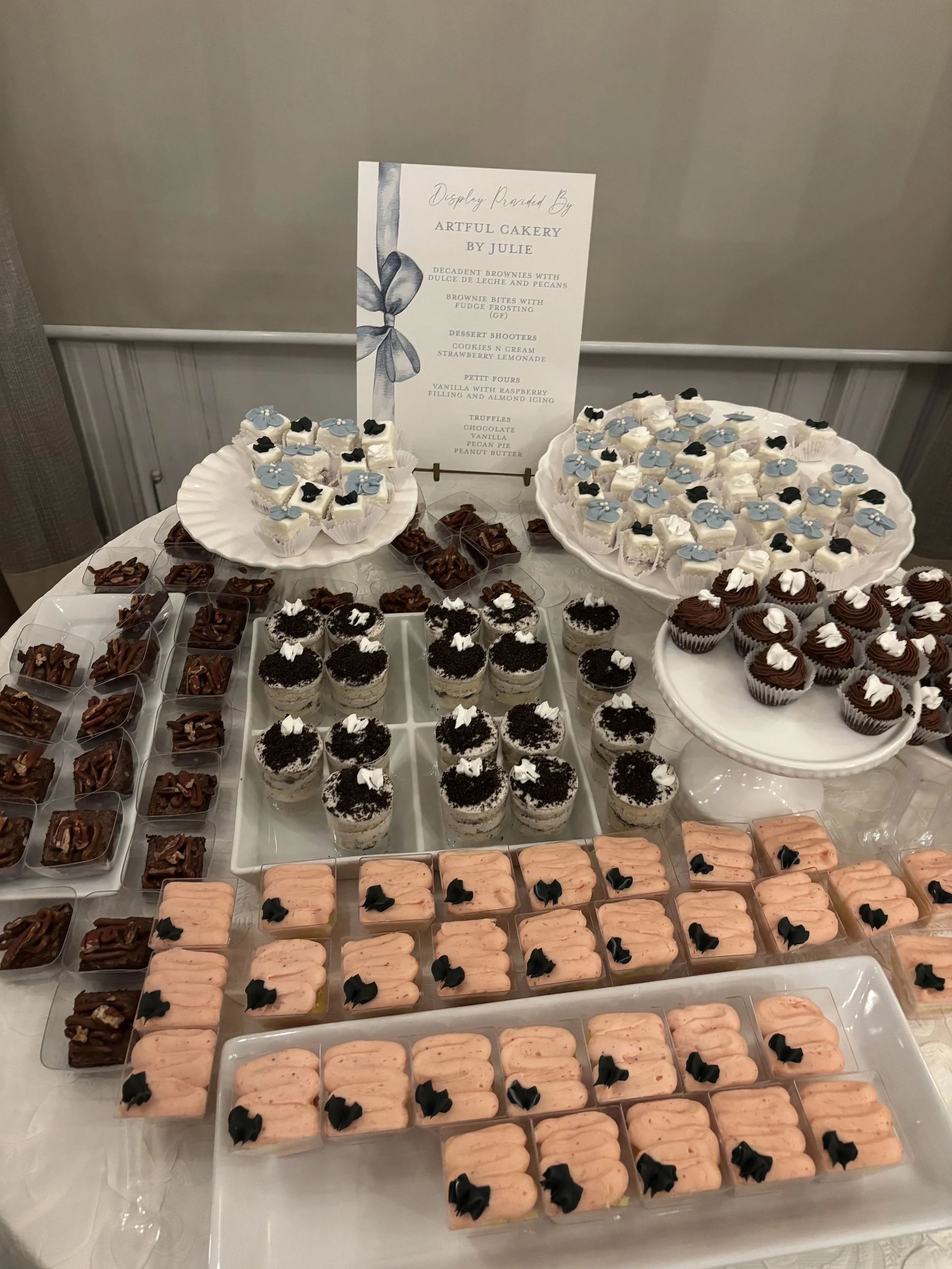 A dessert table with various pastries and cupcakes, including brownies, mini cheesecakes topped with Oreo crumbs, chocolate cupcakes with white frosting, peach-flavored bars with black decorative toppers, and a menu sign listing dessert options.