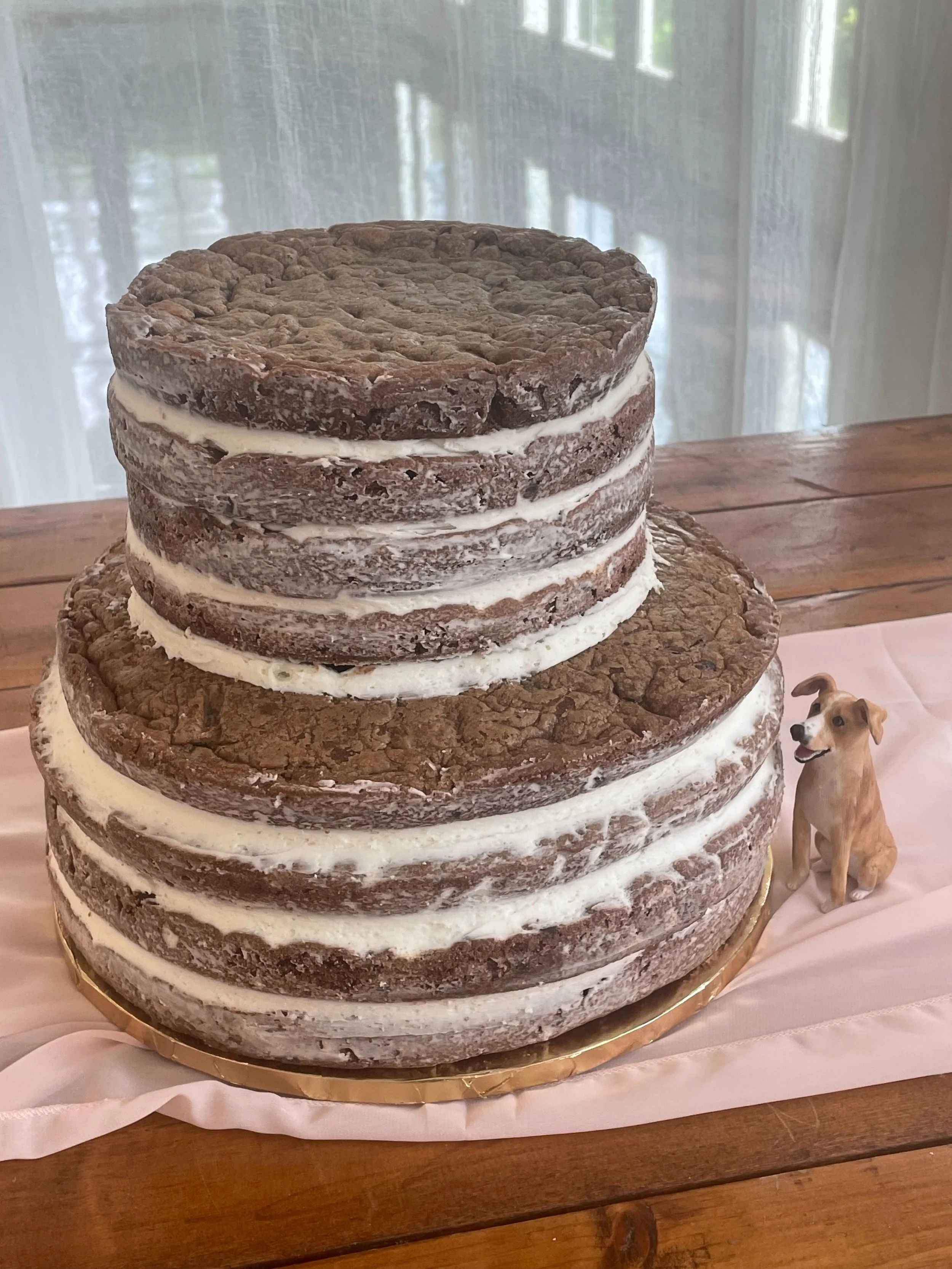 A large, rustic, three-tiered cake designed to look like giant chocolate chip cookies with white icing in between layers, displayed on a wooden table with a small dog figurine beside it.