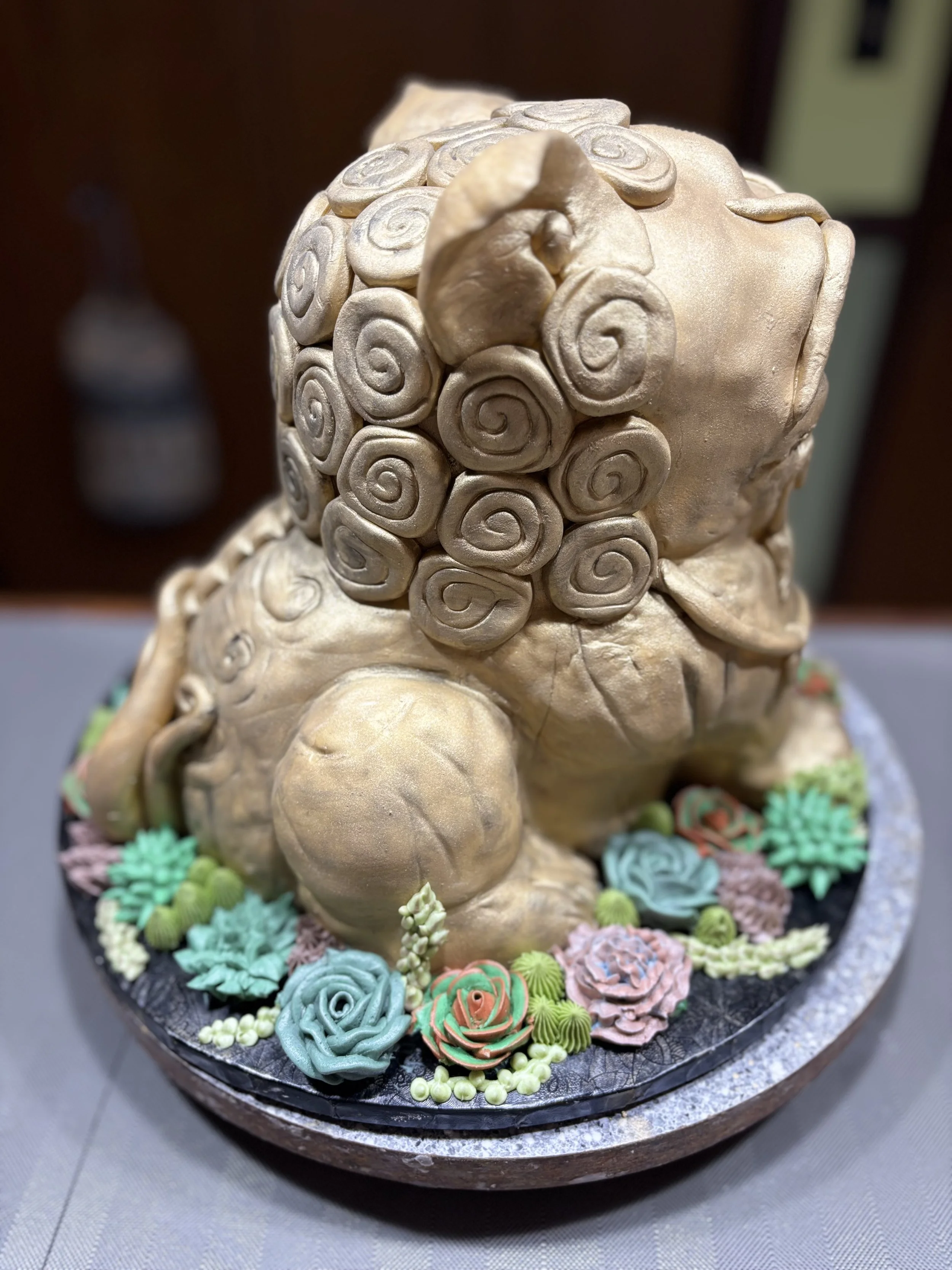 Sculpted cake resembling a Chinese foo dog, with a detailed curly fur texture, surrounded by colorful buttercream flowers and greenery on a cake board.