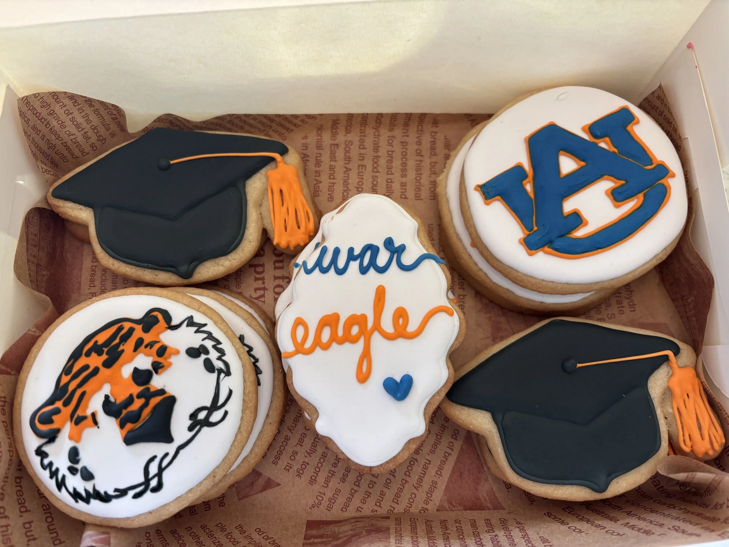 Decorated cookies with graduation theme, including a black cap with an orange tassel, a "War eagle" cookie, an Auburn University logo cookie, and a cookie featuring a tiger's face with black, orange, and white icing.