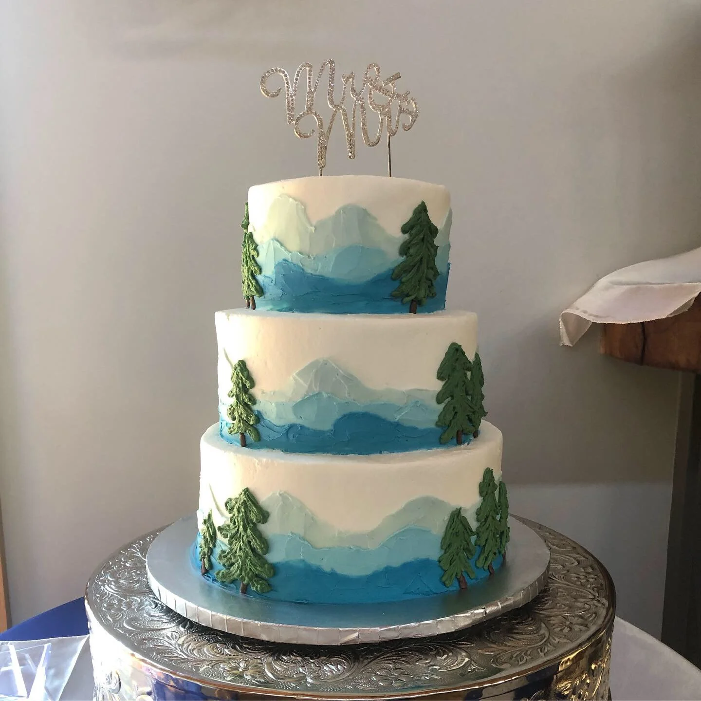 Three-tier wedding cake featuring blue mountain landscapes and textured buttercream evergreen trees at Juliette Chapel in Dahlonega, Georgia.