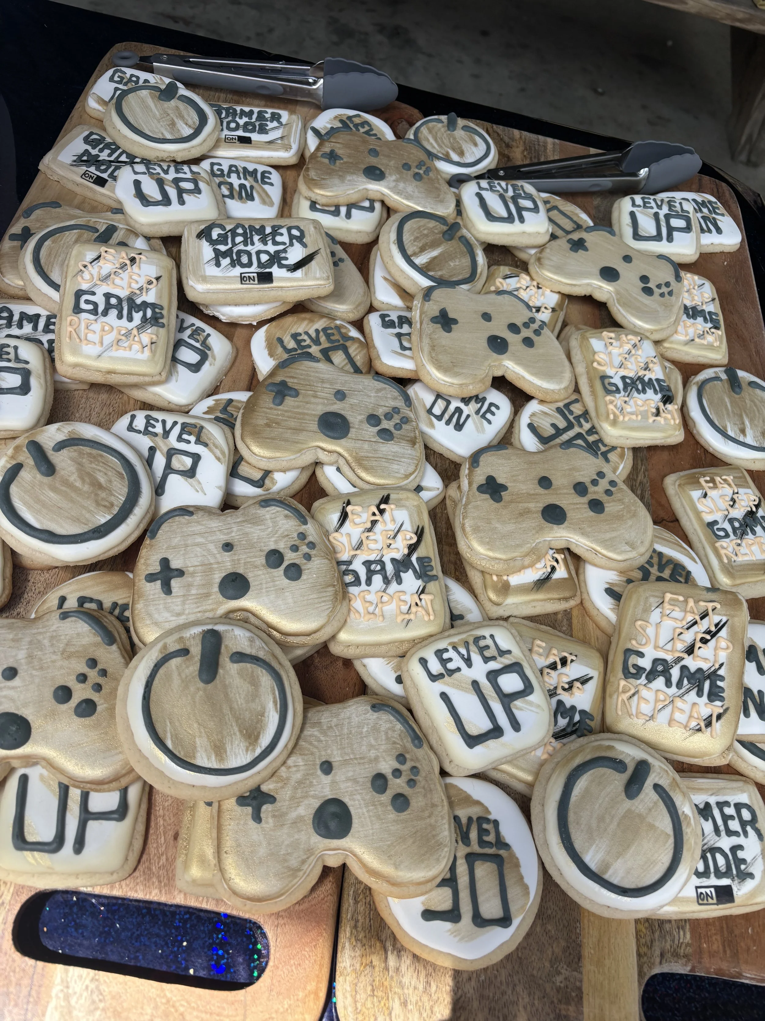 Cookies decorated with gaming themes, including phrases like 'Level Up,' 'Game On,' 'Eat Sleep Game Repeat,' and controller and power button designs, displayed on a wooden tray.