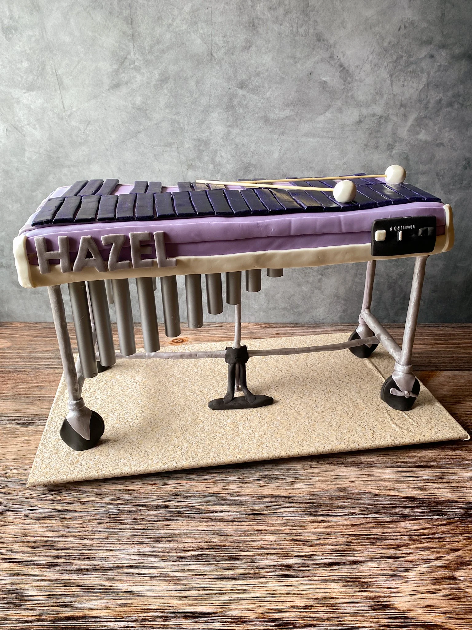 Cake designed to look like a marimba with the name 'Hazel' on the side, placed on a wooden surface against a gray wall.