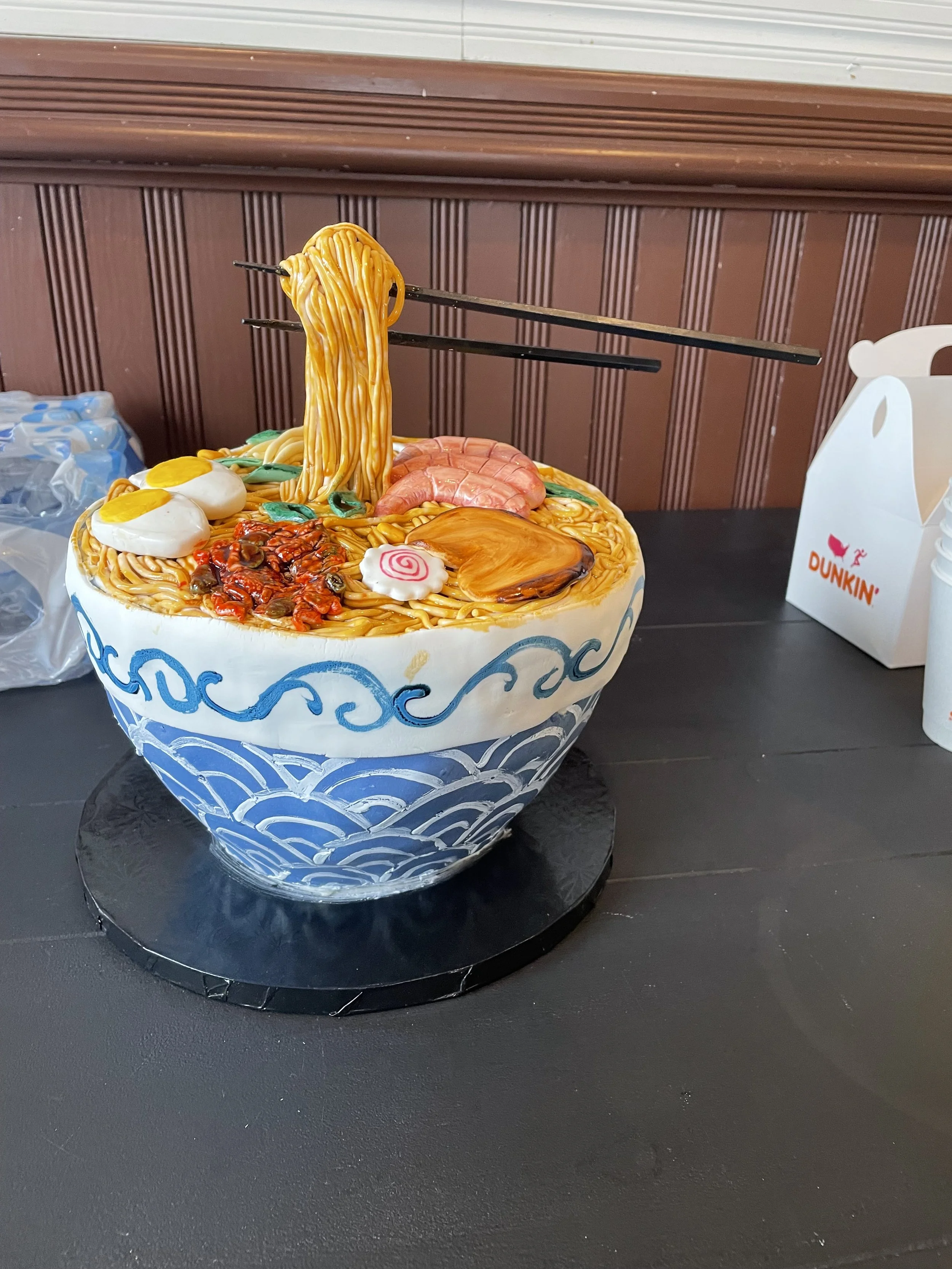 A cake designed to look like a bowl of ramen, with ramen toppings and noodles, on a black surface with a dark wooden wall in the background.