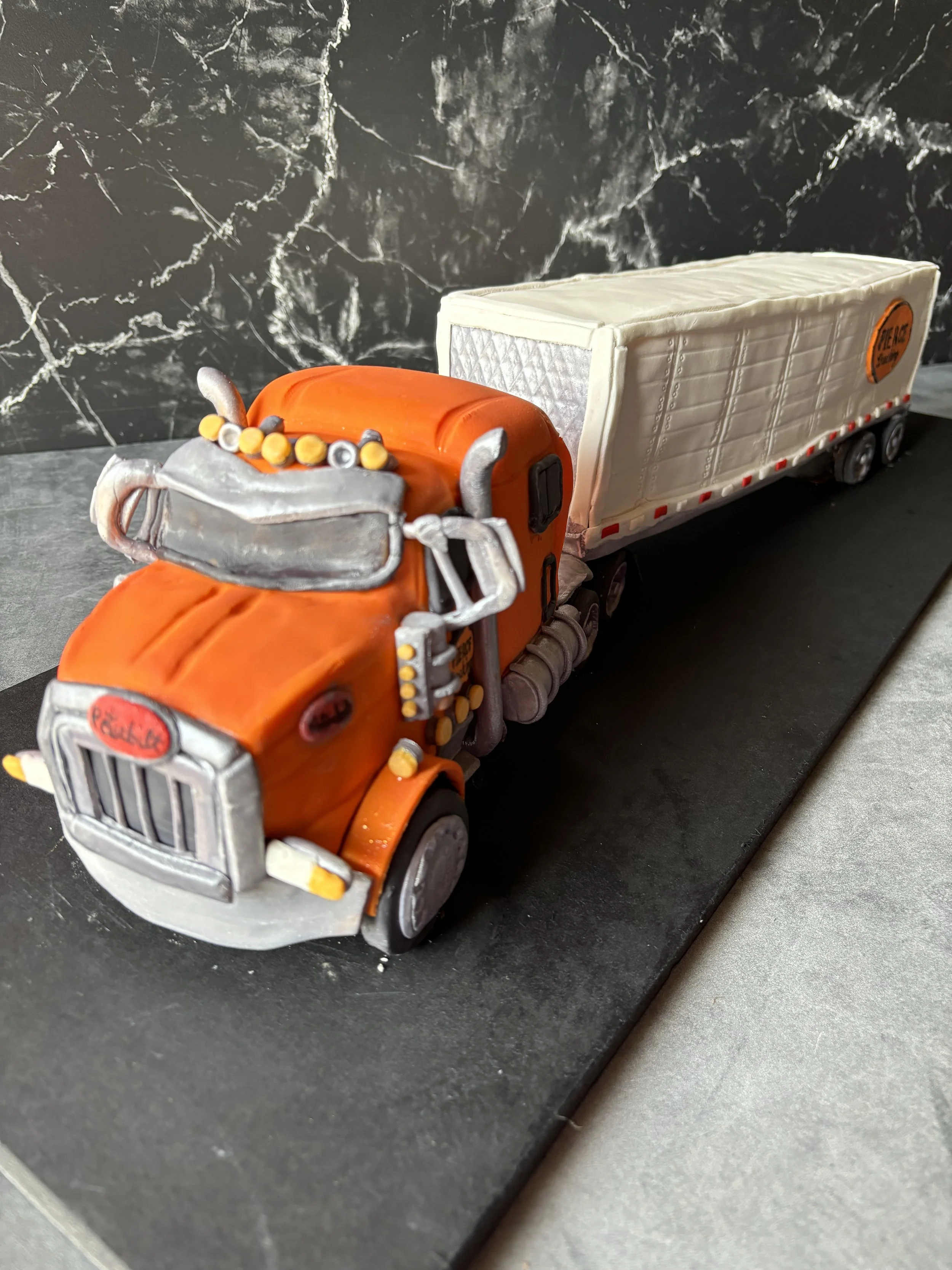 Cake shaped like a semi-truck, with an orange cab and white trailer, on a black surface.