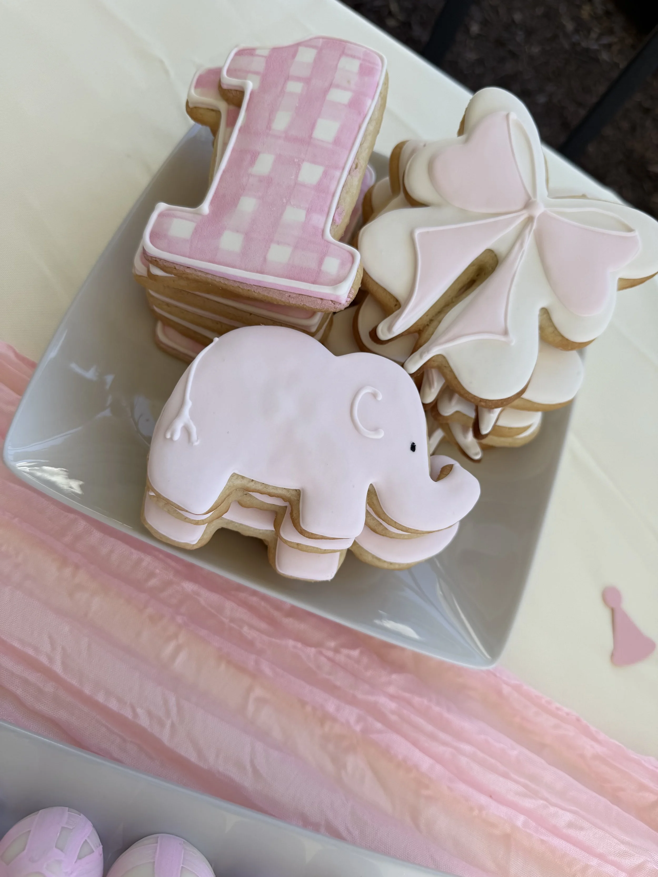 Decorated cookies on a white tray, including pink and white checkered number '1' cookie, white elephant-shaped cookie, and a swan-shaped cookie with pink and white icing, set on a pink tablecloth.